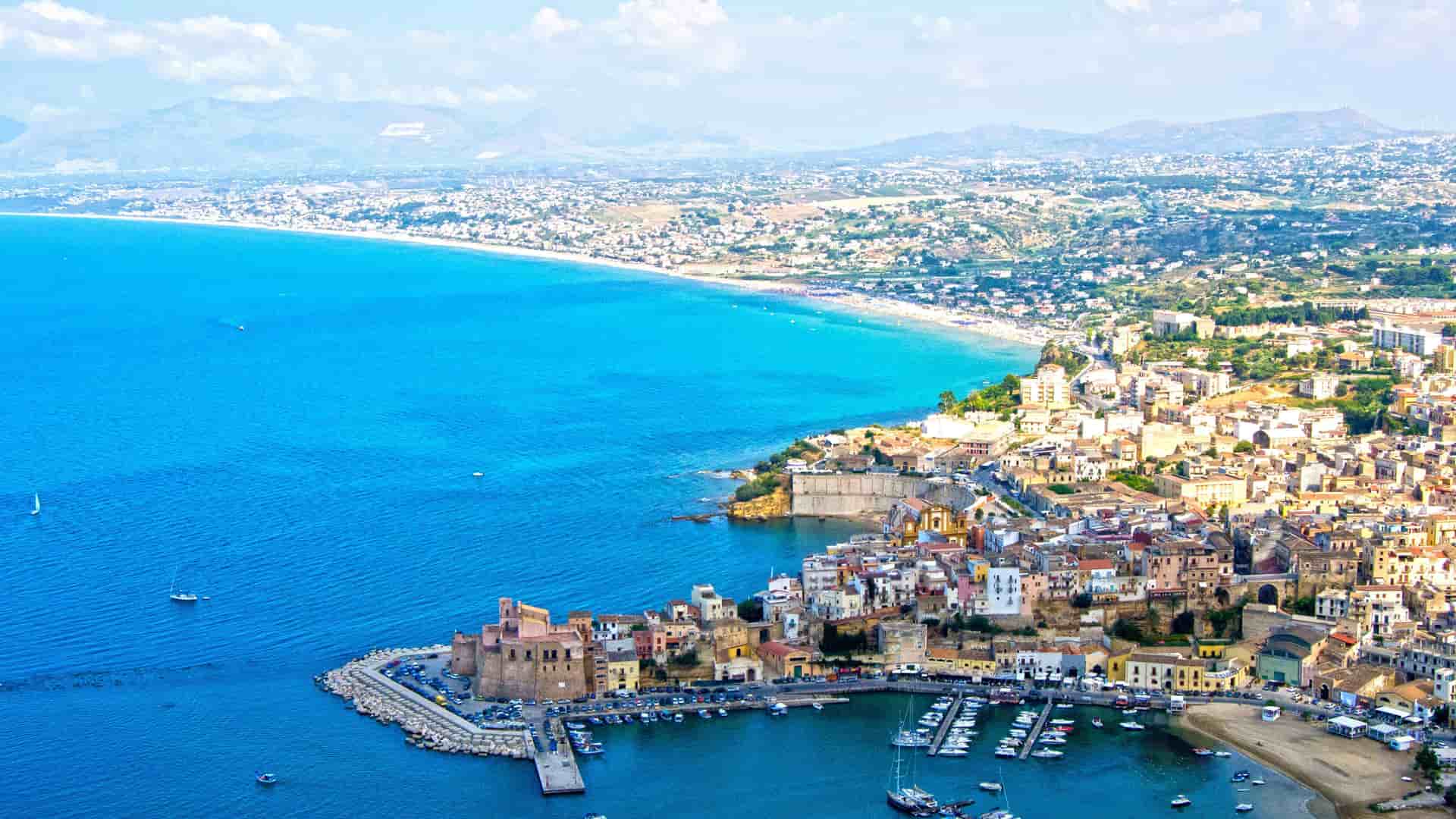 An aerial view of the coastal city of Trapani, Sicily, with its scenic harbor filled with boats and a long sandy beach along the turquoise blue water.
