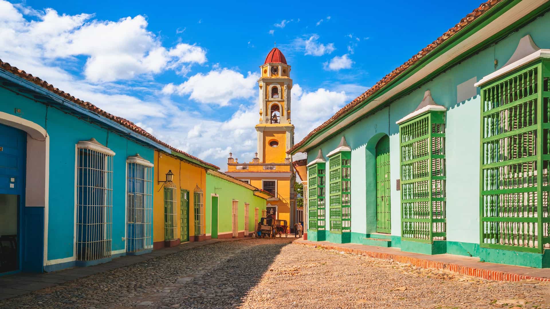 "A cobblestone street in Trinidad, Cuba, lined with colorful historic buildings in shades of blue, green, and yellow, leading towards the iconic bell tower of the Iglesia y Convento de San Francisco. "