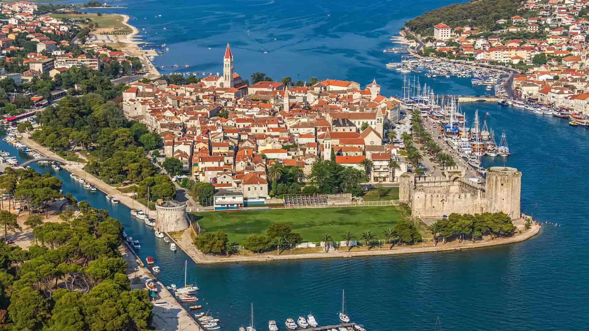 An aerial shot of the historic walled island city of Trogir, Croatia, showing its red-roofed buildings, marina filled with yachts, and a medieval fortress surrounded by blue water.