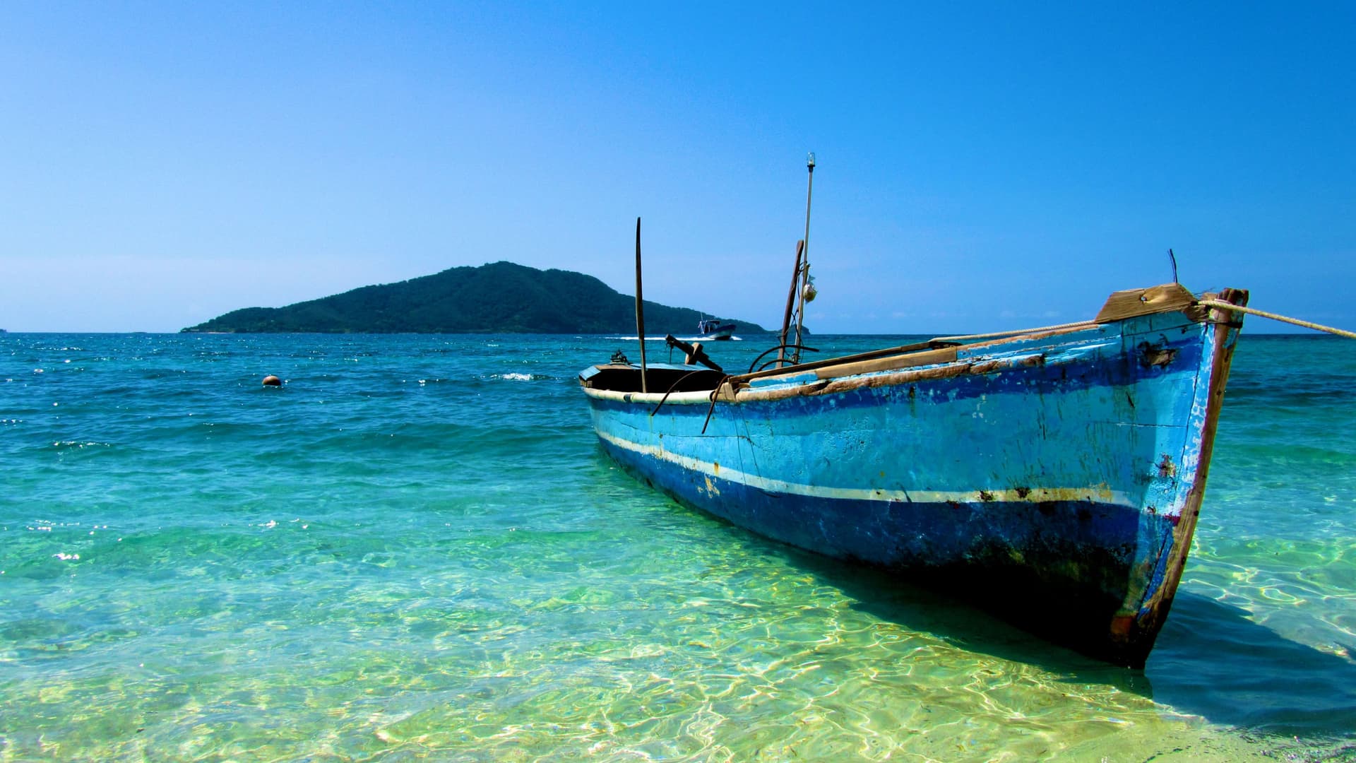 A blue, rustic fishing boat floats in the turquoise and clear water of a tropical beach near Trujillo, Peru, with a small, green island in the distance.