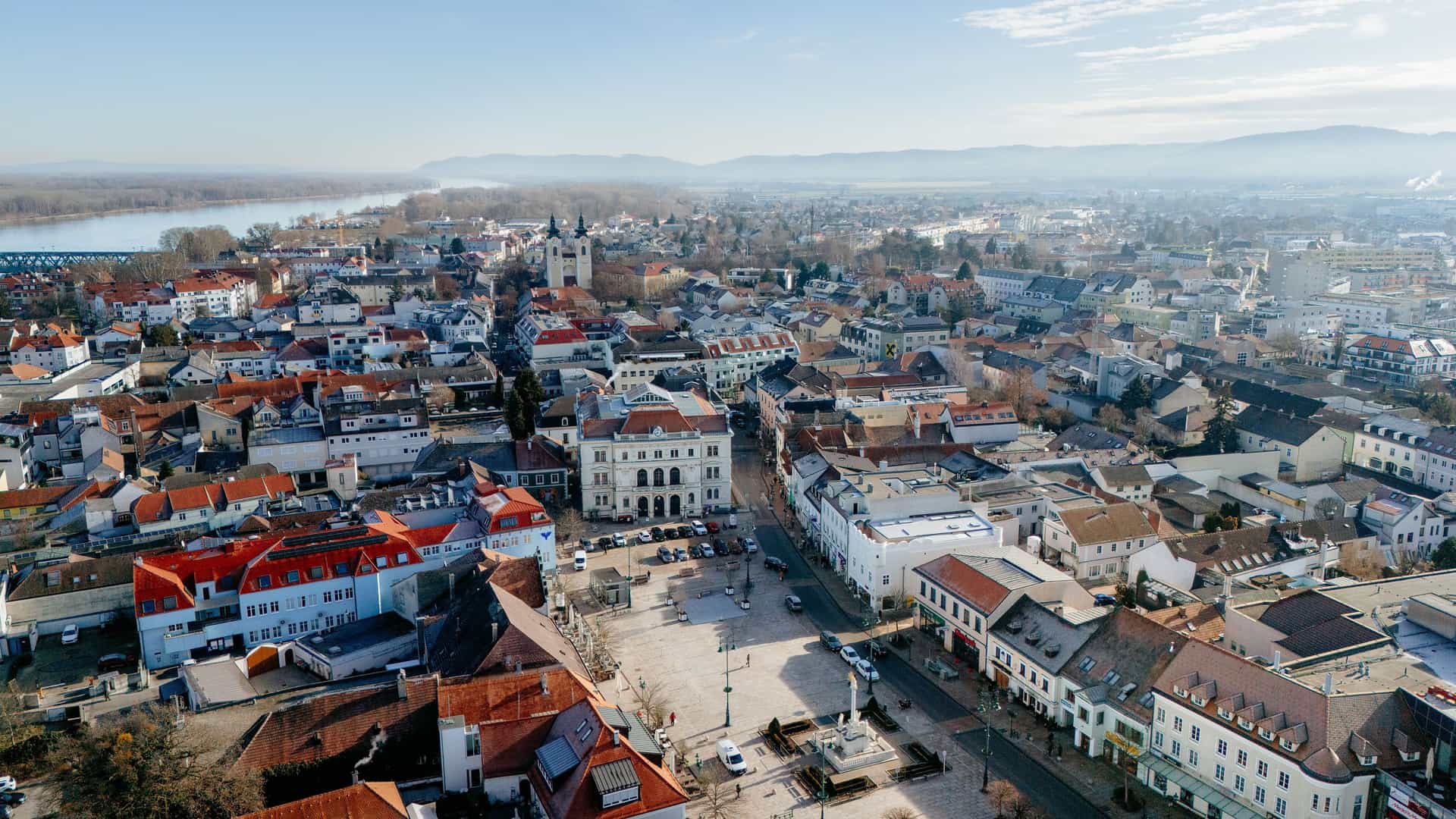 An aerial view of the historic town of Tulln an der Donau, Austria, showing the main square with buildings and a view of the Danube River and surrounding mountains.