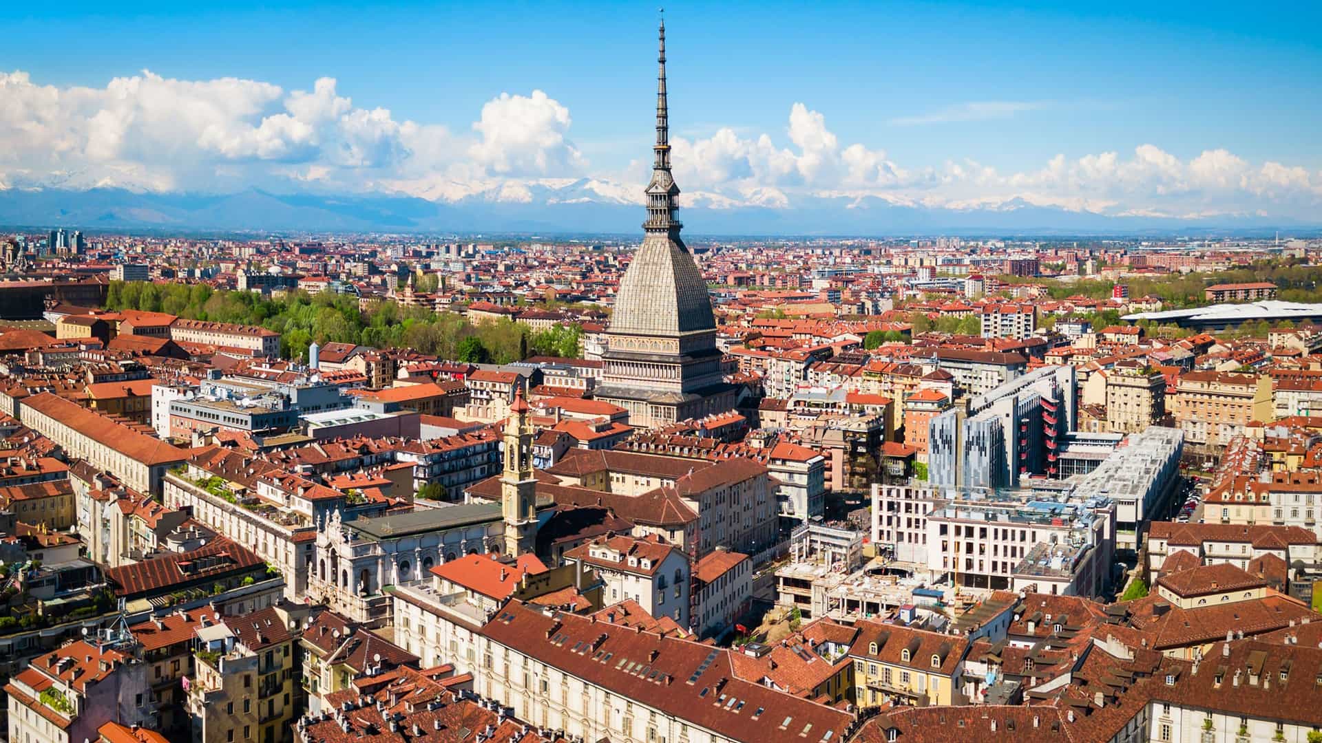 "An aerial panoramic shot of Turin, Italy, highlighting the iconic Mole Antonelliana spire rising above the cityscape of red-tiled roofs, with the snow-capped Alps in the background. "