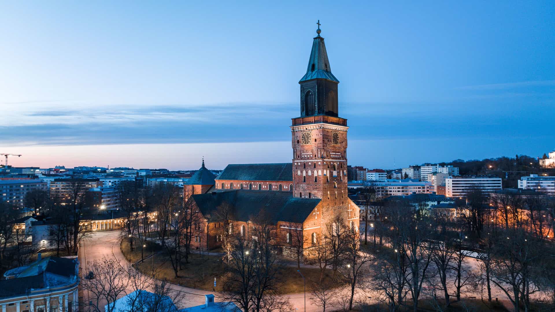 An aerial shot of the medieval Turku Cathedral illuminated at dusk, its brick exterior and tall spire a prominent landmark in the Finnish cityscape.