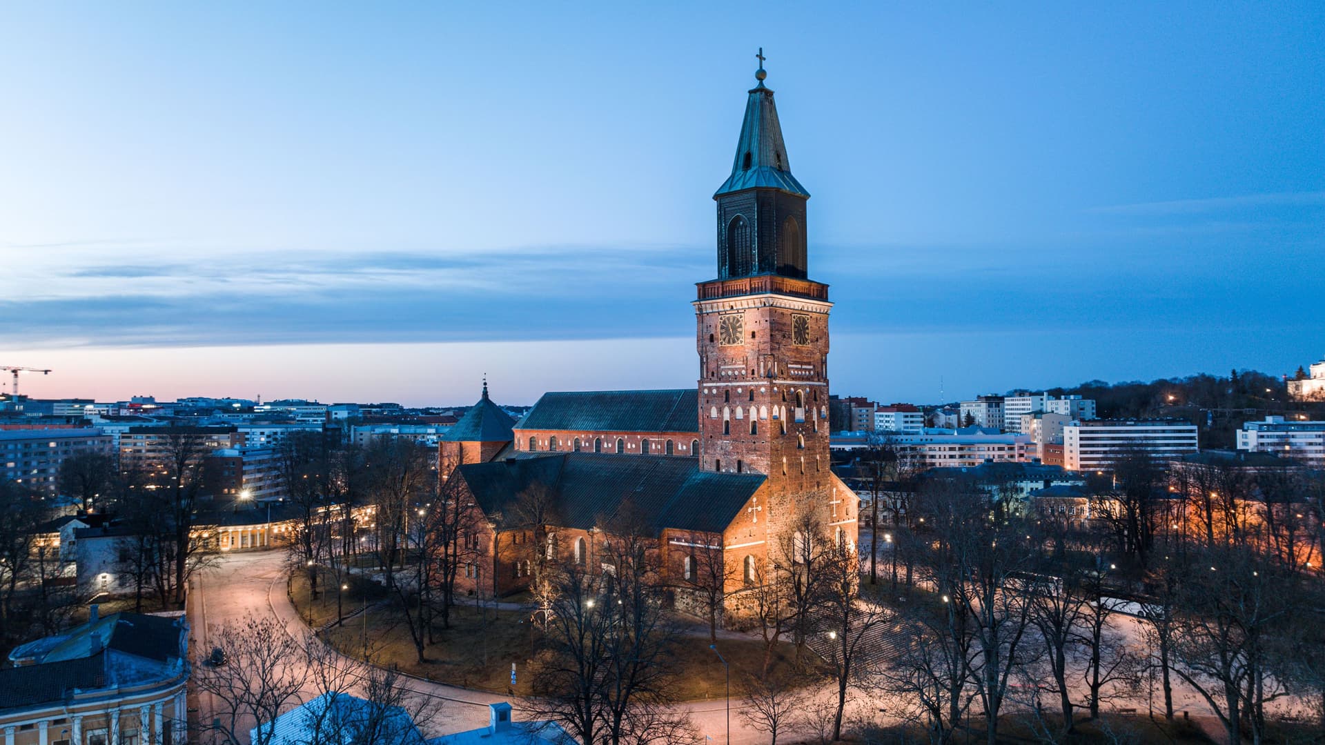 An aerial shot of the medieval Turku Cathedral illuminated at dusk, its brick exterior and tall spire a prominent landmark in the Finnish cityscape.