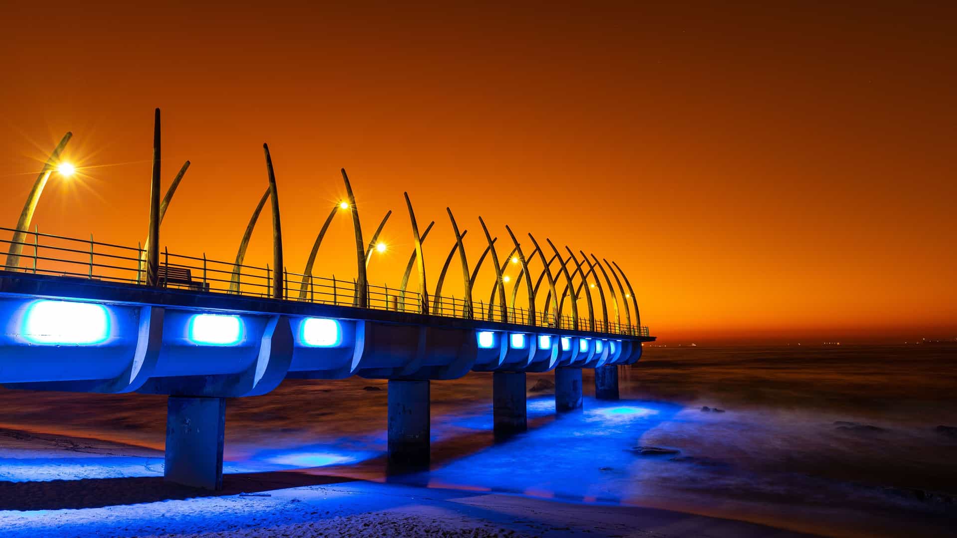 "A striking nighttime view of the Whalebone Pier in Umhlanga, South Africa, illuminated with bright blue lights, with the orange glow of the rising sun or the city lights reflecting off the night sky. "
