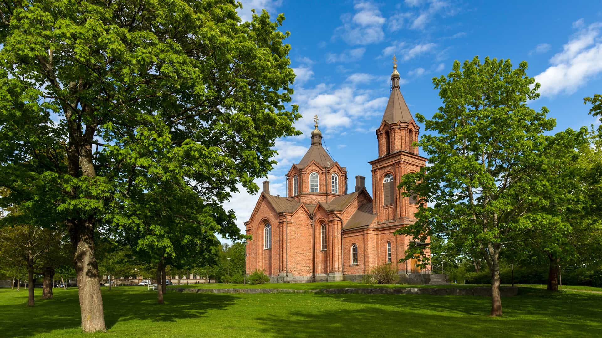 A picturesque view of the red brick Vaasa Orthodox Church with its golden domes, surrounded by a lush green park with trees and grass under a bright blue sky.