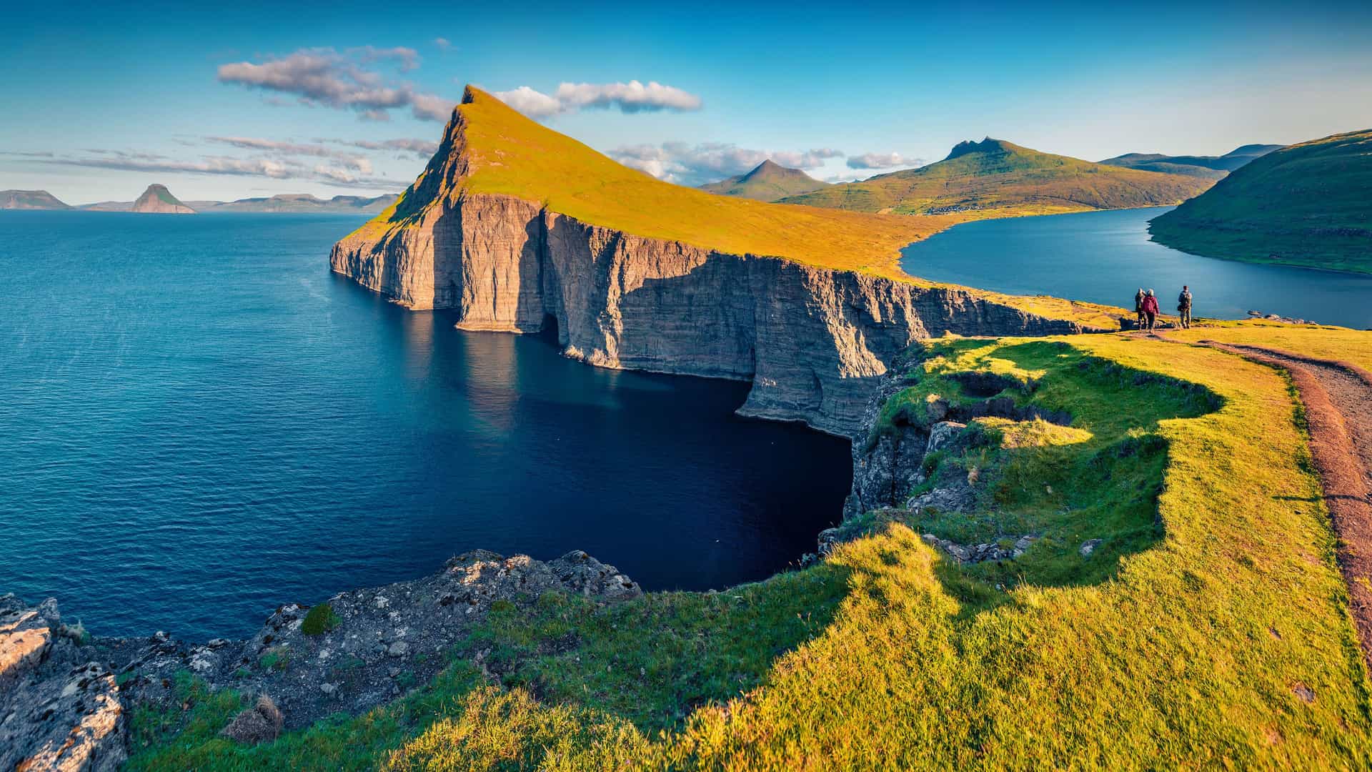 "A breathtaking panoramic shot of Lake Sørvágsvatn, also known as ""The Lake Above the Ocean,"" on the island of Vágar in the Faroe Islands, Denmark. The image shows the steep, grassy cliffs and calm water with a hiking trail and a person in the foreground. "