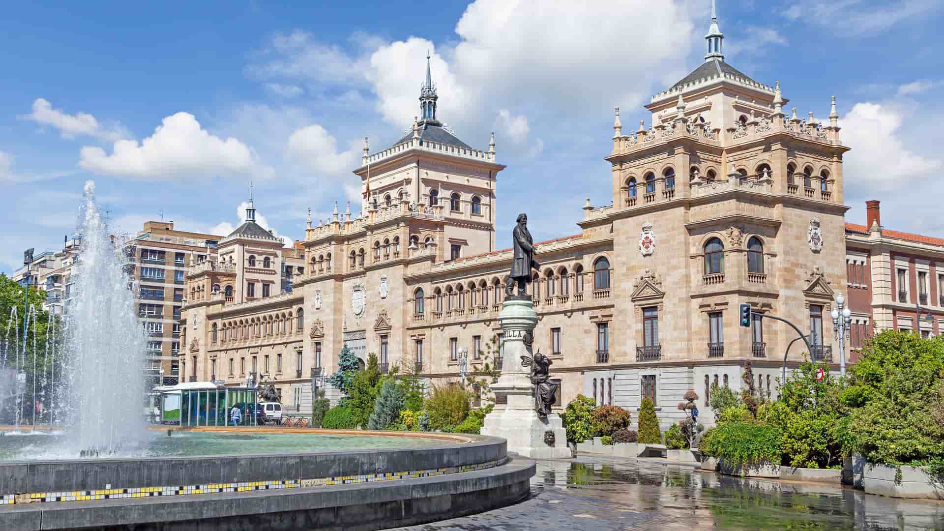 "A sunny daytime shot of the Royal Palace of Valladolid, Spain, with a large fountain in the foreground and a bronze statue in front of the ornate, historic brick building. "