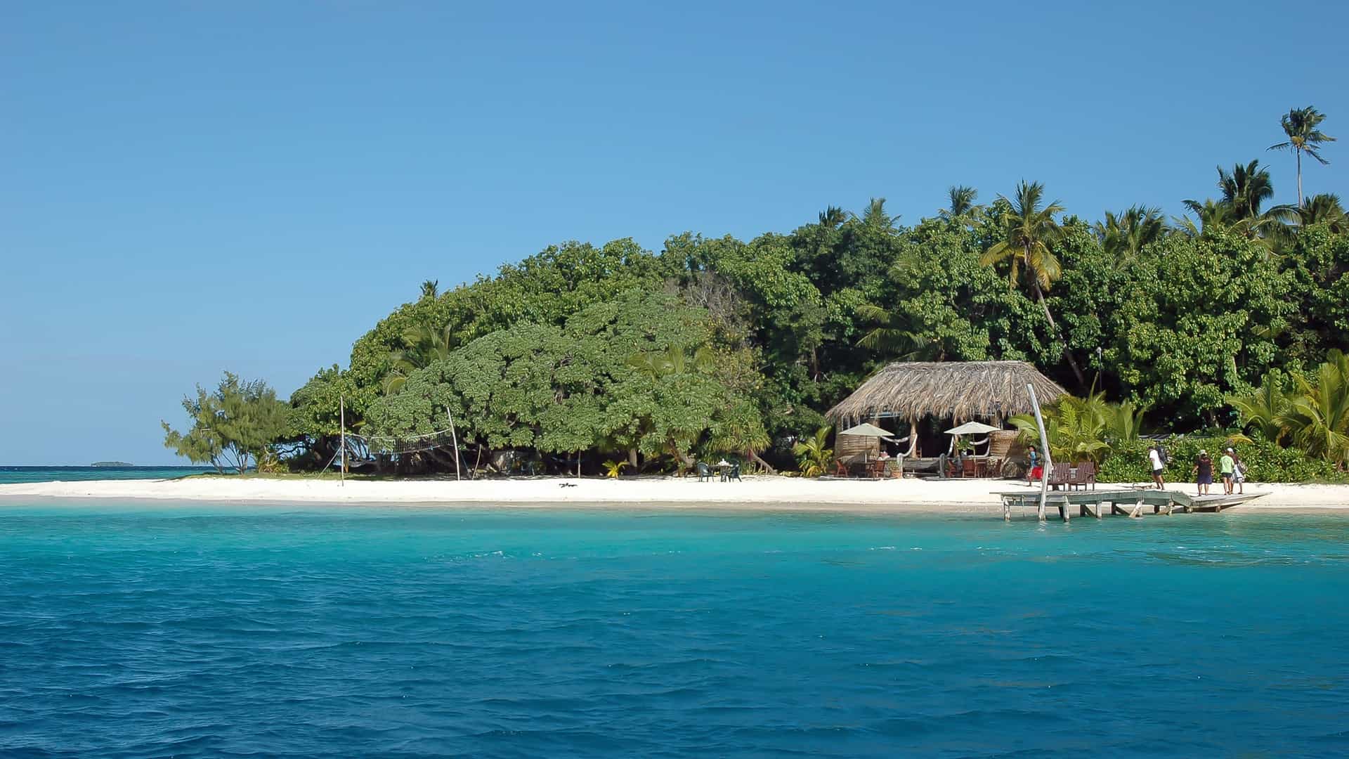 A pristine white-sand beach with a rustic thatched-roof hut on the coast of a lush, tropical island in Vavaʻu, Tonga, with people standing on a small wooden pier.