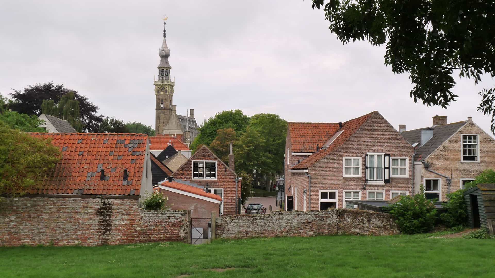 A row of historic red brick houses with red tiled roofs, a stone wall, and a church steeple in the background in the quaint town of Veere, Netherlands.