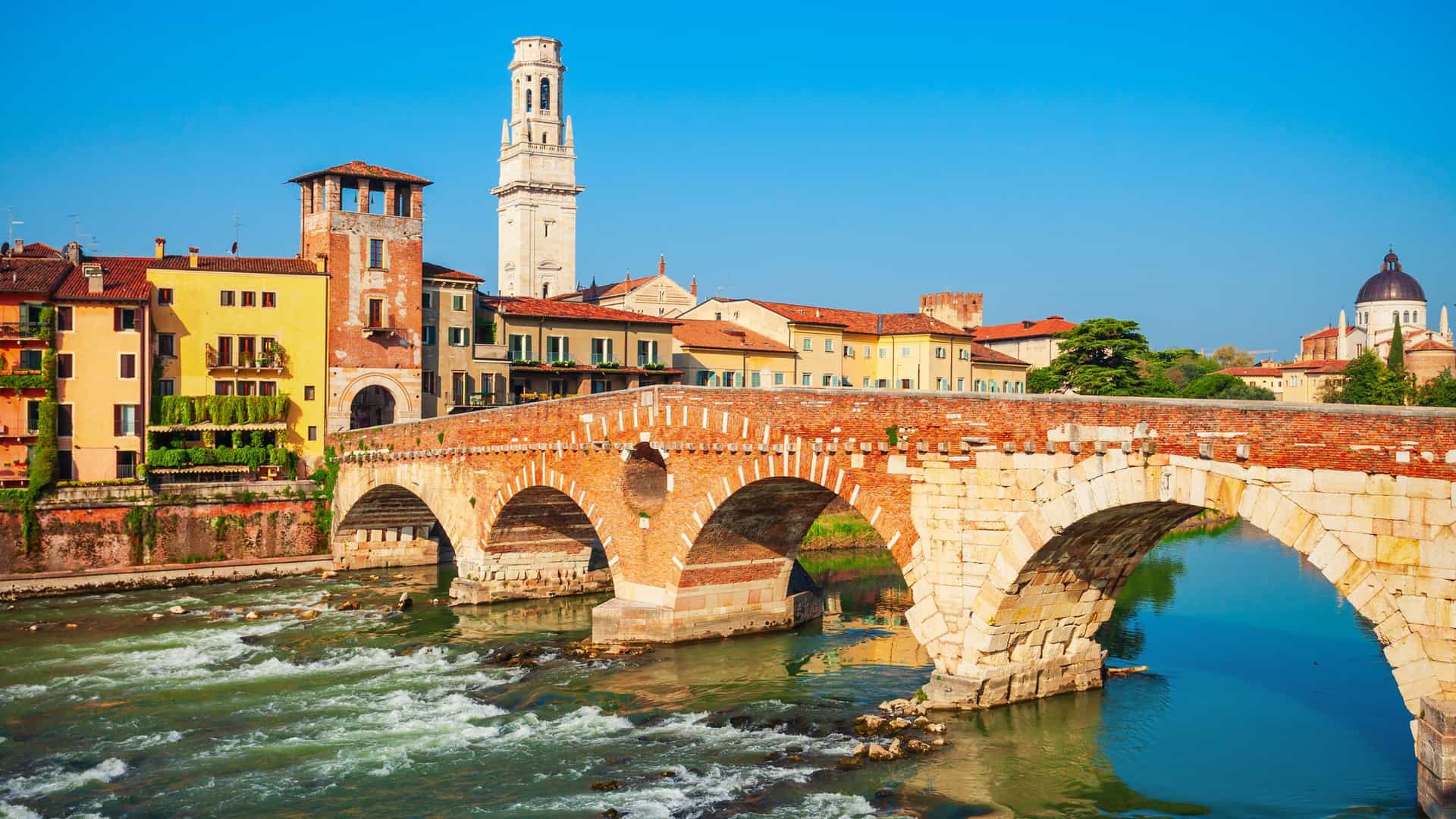 A sunny view of the historic Ponte Pietra bridge over the Adige River in Verona, Italy, with the medieval city buildings and the bell tower of the Verona Cathedral in the background.