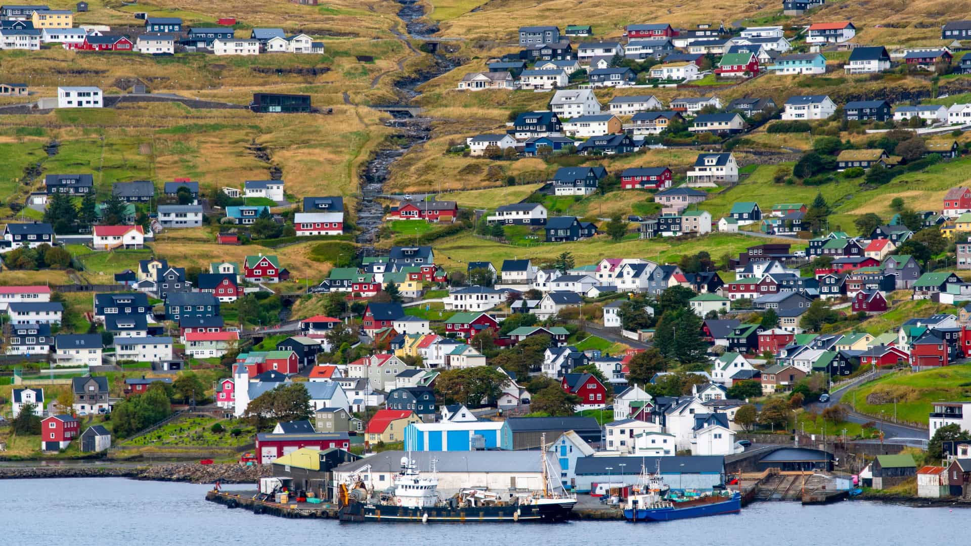 A beautiful view of the coastal town of Vestmanna, Faroe Islands, with its colorful houses scattered on a steep green hillside overlooking a small harbor.