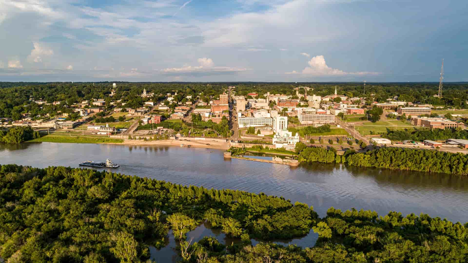 An aerial view of the historic city of Vicksburg, Mississippi, with a barge on the expansive Mississippi River, surrounded by lush green trees under a partly cloudy sky.