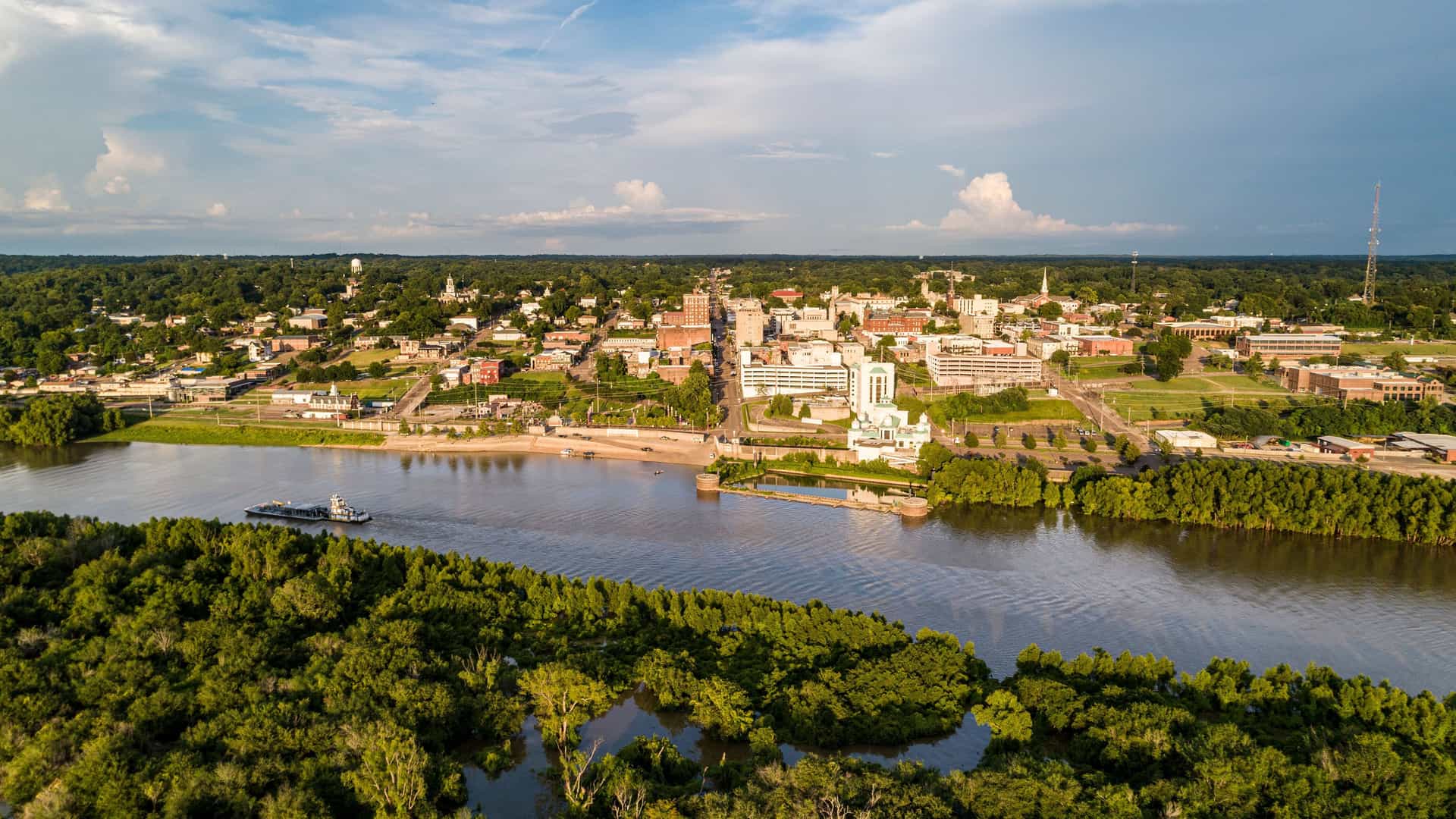 An aerial view of the historic city of Vicksburg, Mississippi, with a barge on the expansive Mississippi River, surrounded by lush green trees under a partly cloudy sky.