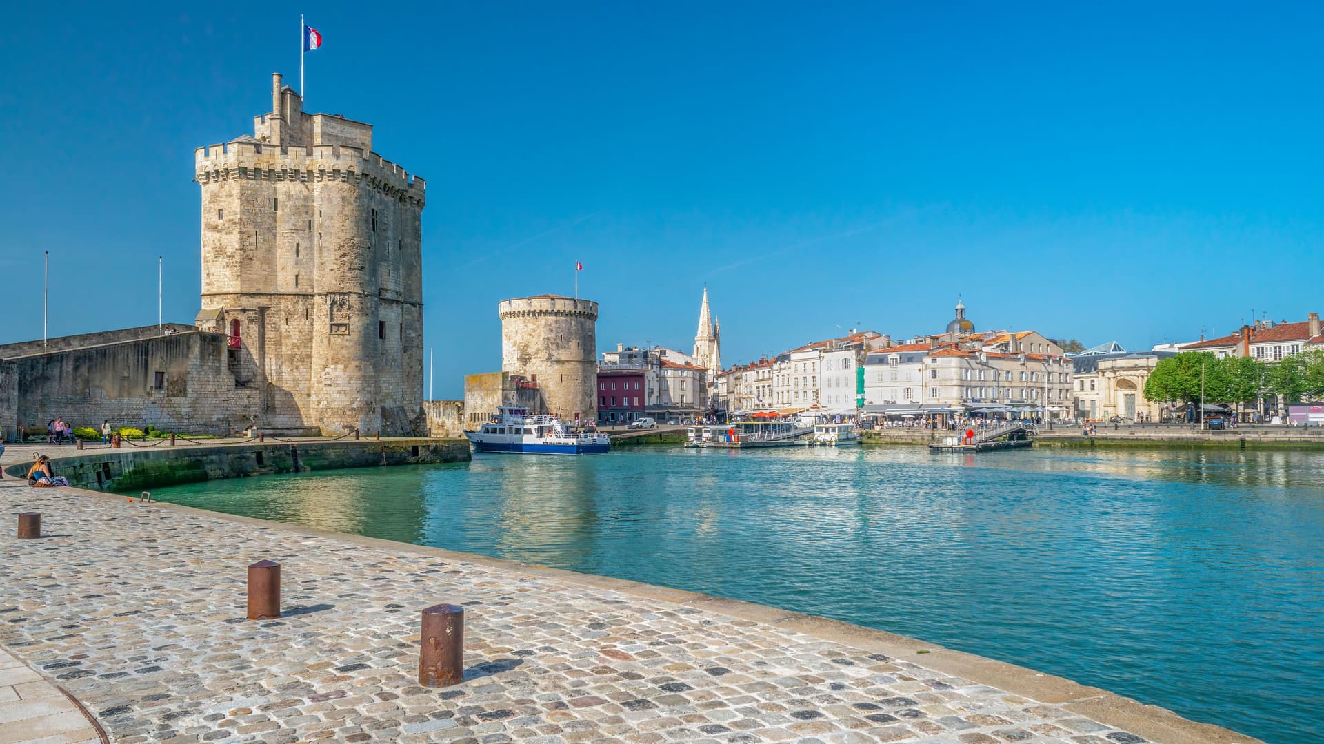 A vibrant harborside view of the historic medieval Towers of La Rochelle, a major landmark, with a cobblestone quay and boats floating in the calm water on a sunny day.