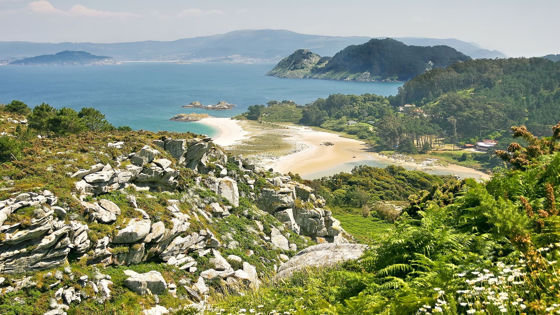 A panoramic high-angle view of the scenic Cíes Islands archipelago in Vigo, Spain, with a sheltered beach and green hills cascading down to the calm blue waters of the bay.
