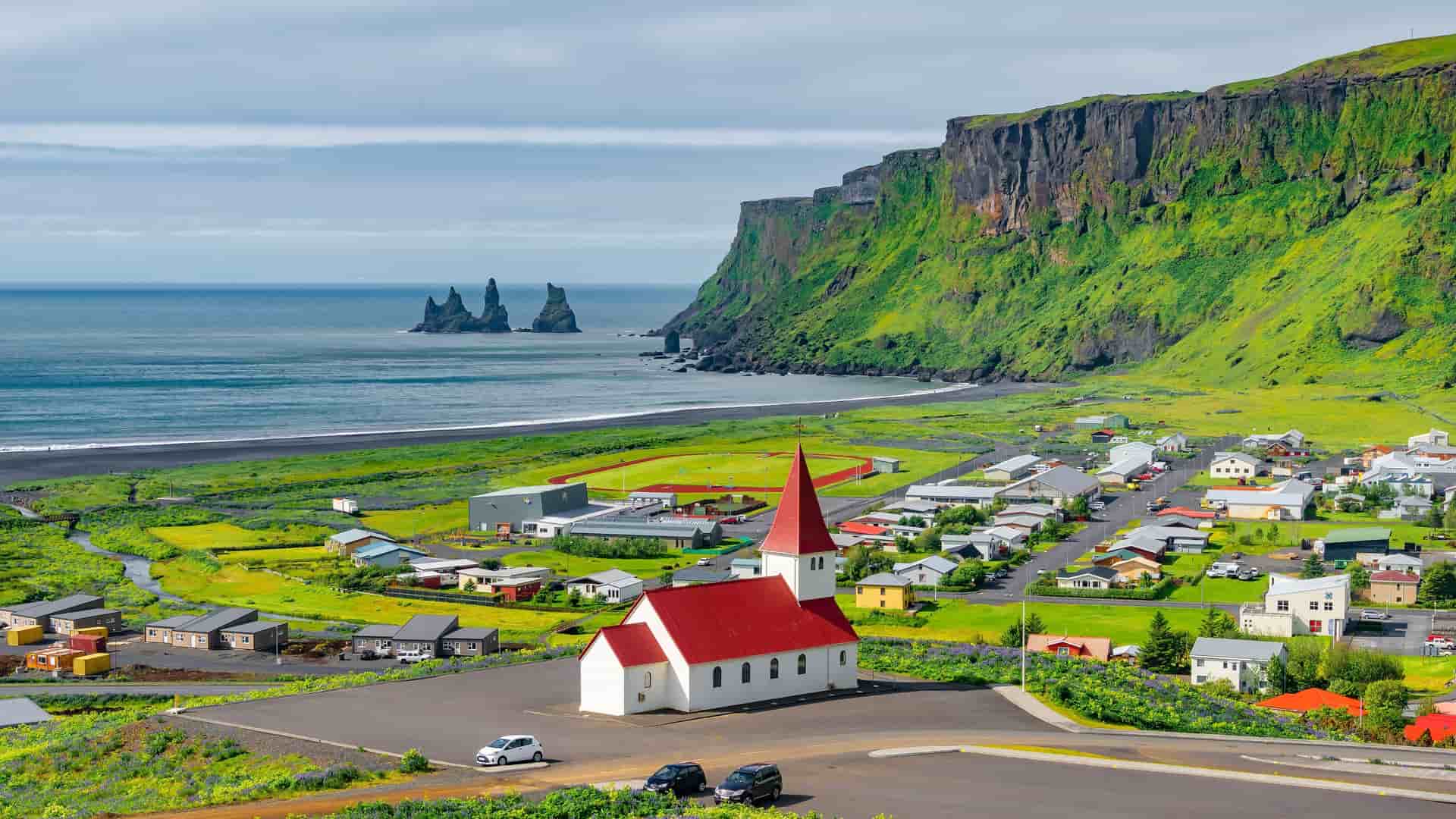 "A high-angle view of the charming seaside village of Vík, Iceland, featuring a classic white church with a bright red roof. The village is nestled between a black sand beach and a lush green cliff, with the famous Reynisdrangar sea stacks visible offshore. "