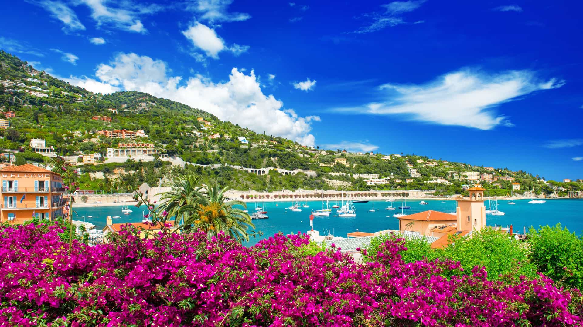 A vibrant view of the picturesque town of Villefranche-sur-Mer, France, with colorful buildings and yachts in the bay, set against a backdrop of steep, green hills under a bright blue sky.