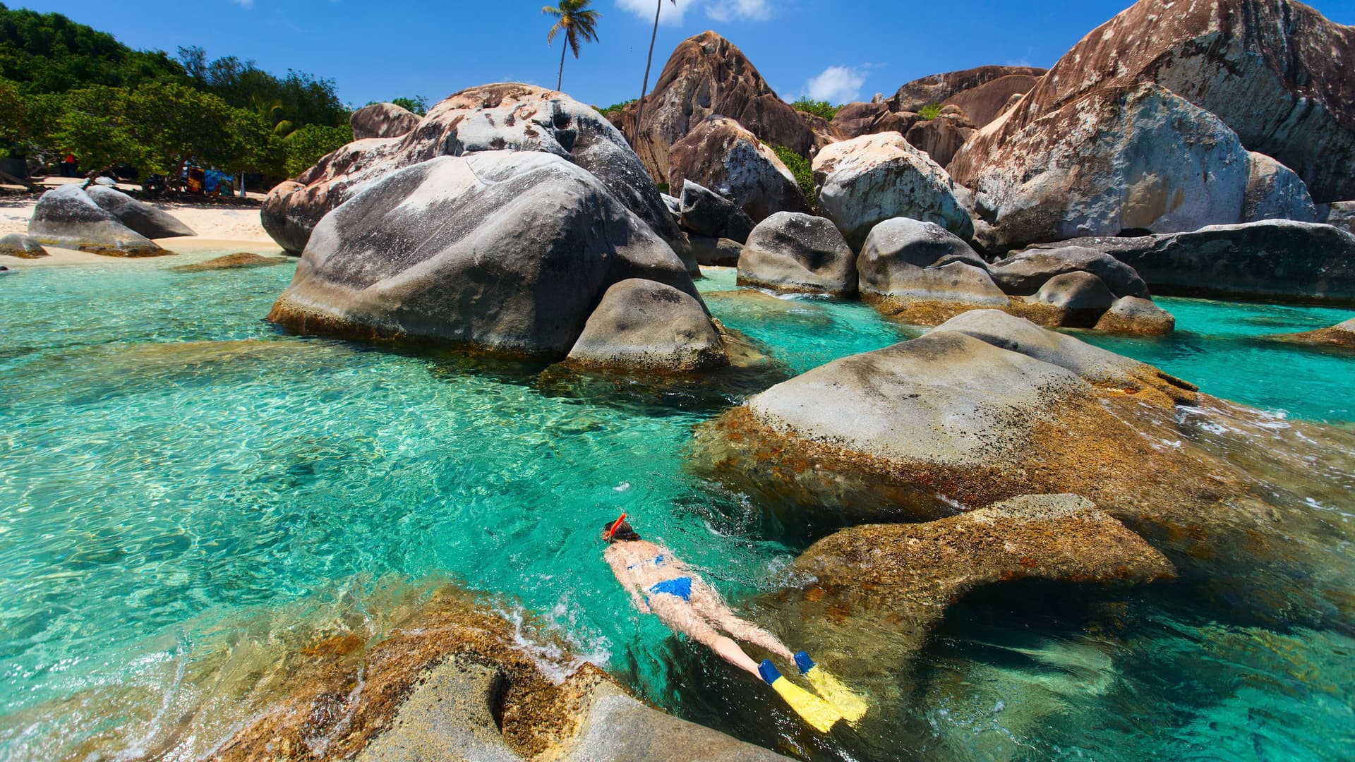A snorkeler swims in the turquoise waters of the Baths National Park on Virgin Gorda, surrounded by massive granite boulders and palm trees.