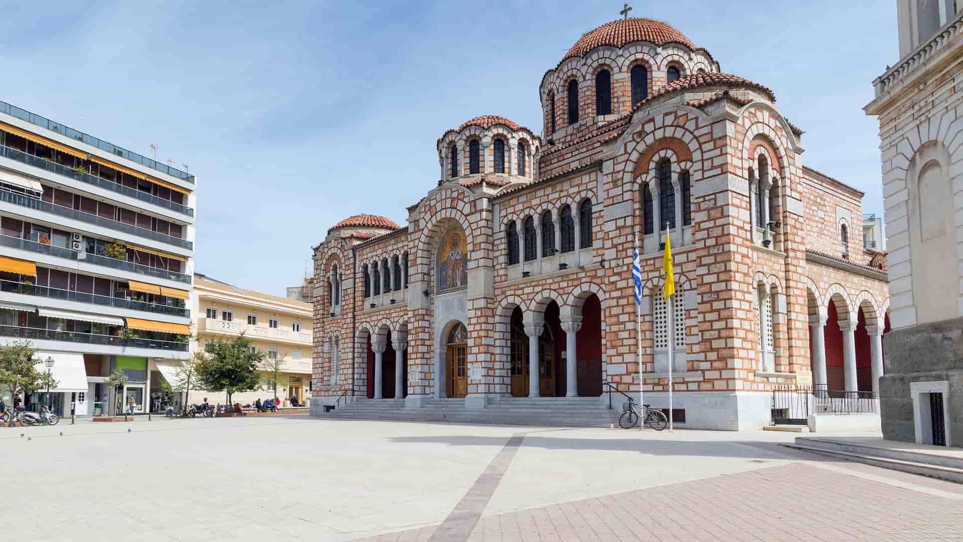 A daytime shot of the Agios Nikolaos church in Volos, Greece, with its distinctive brickwork and domes standing in a large, open square.