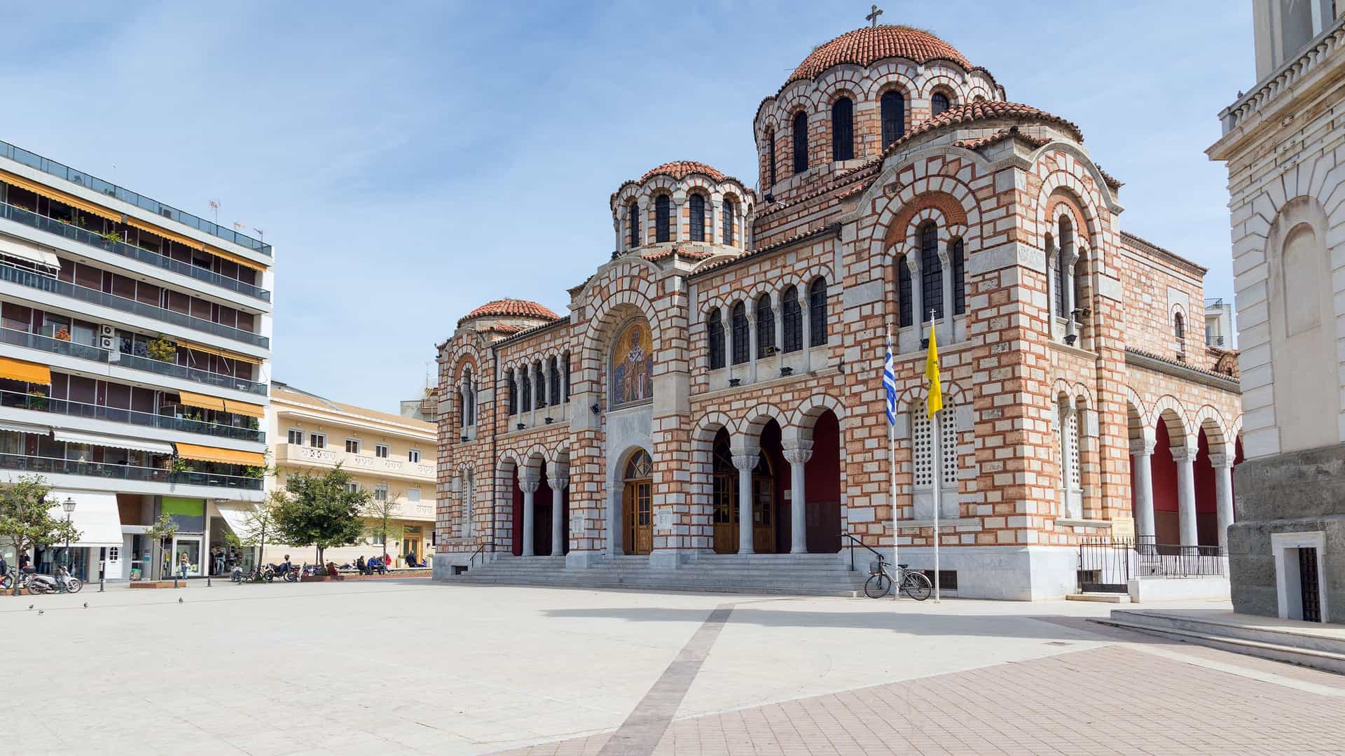 A daytime shot of the Agios Nikolaos church in Volos, Greece, with its distinctive brickwork and domes standing in a large, open square.