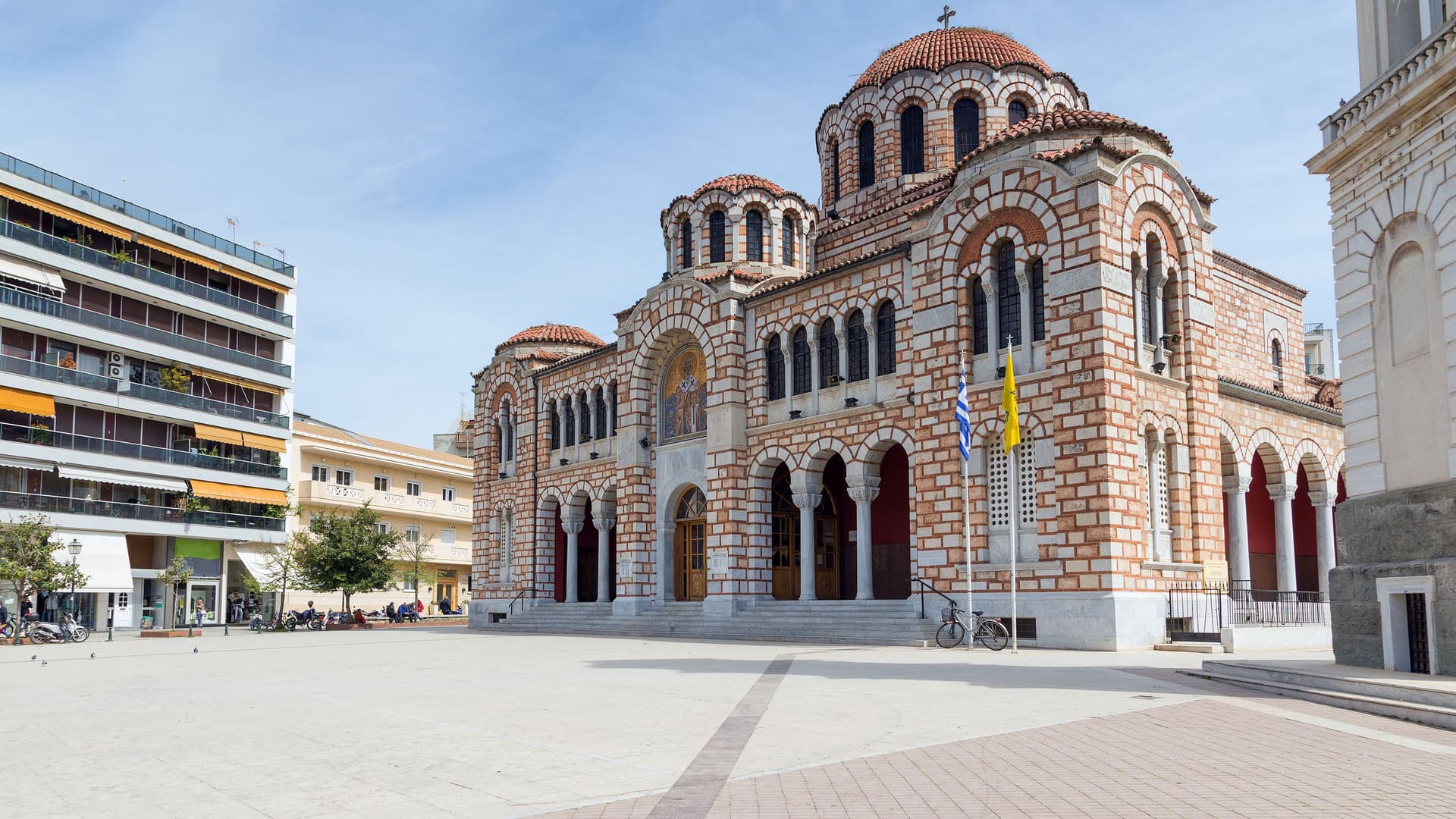 A daytime shot of the Agios Nikolaos church in Volos, Greece, with its distinctive brickwork and domes standing in a large, open square.
