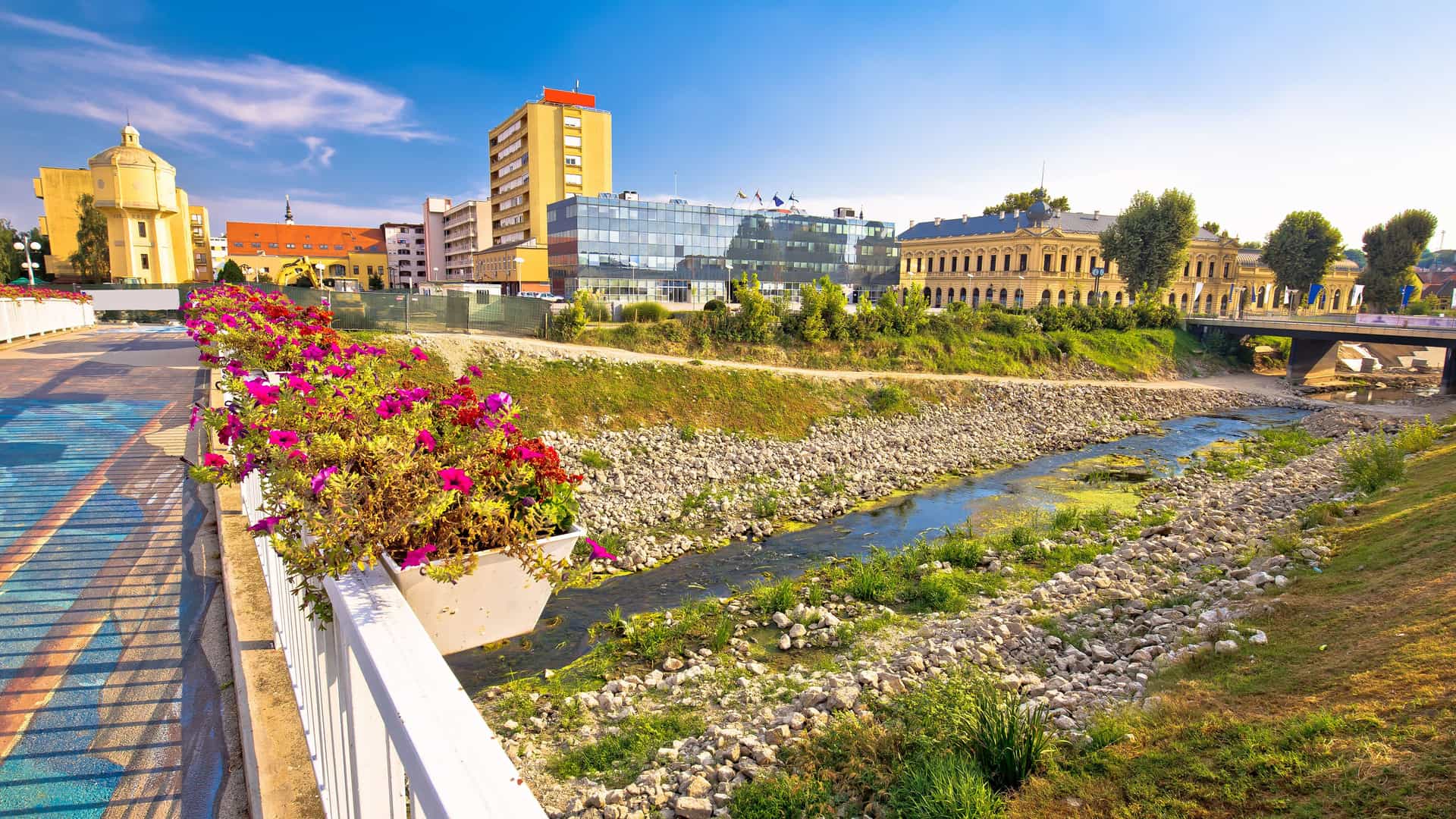 A vibrant riverfront view in Vukovar, Croatia, showcasing the old and modern cityscape with the Duna River, colorful flowers on a bridge, and a mix of historic and new buildings in the background.