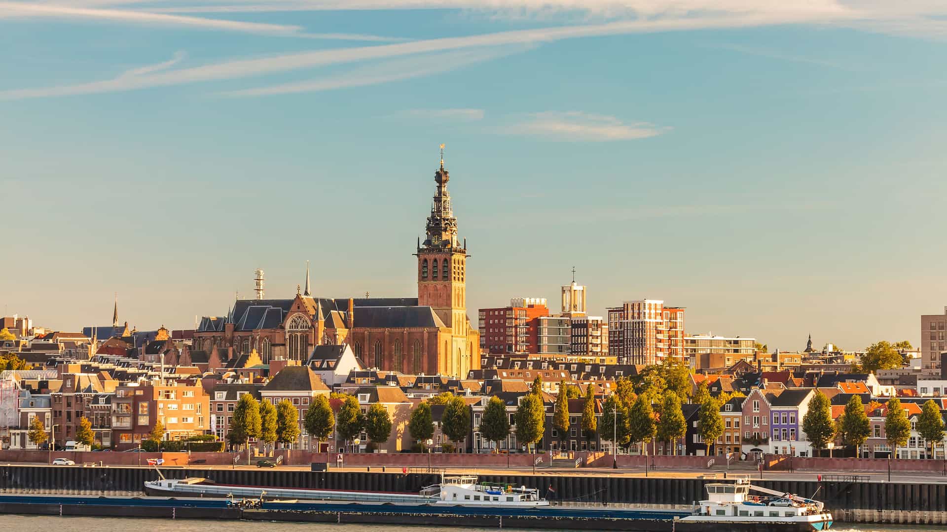 A panoramic view of the historic city of Nijmegen on the Waal and Merwede rivers in the Netherlands, with the prominent tower of the Grote Kerk church overlooking the bustling waterway with boats.