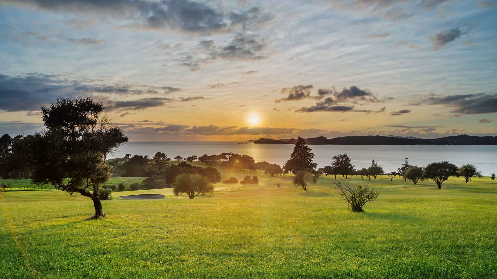 A serene sunset view over the Bay of Islands in Waitangi, New Zealand, with the sun setting over the water, casting a golden glow on the green landscape.