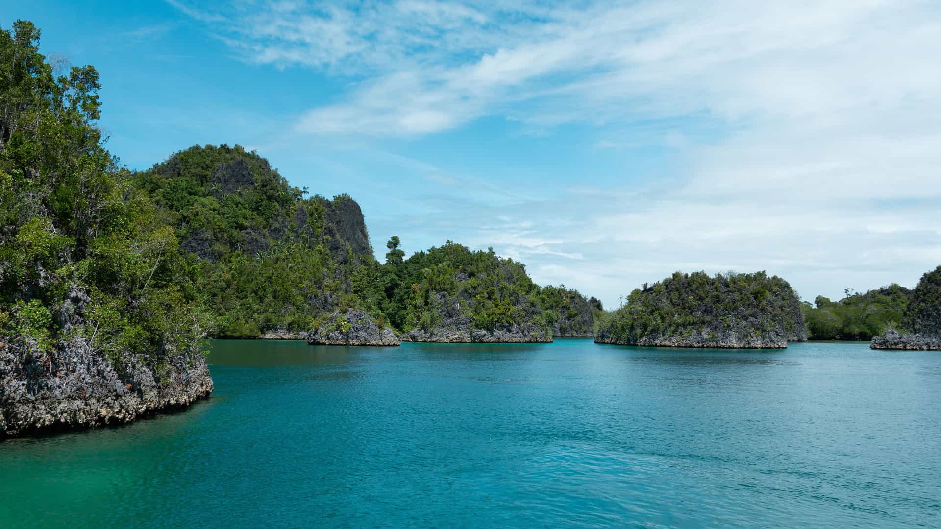A stunning view of the lush, green limestone islands of Wajag Island, Indonesia, jutting out of the calm, turquoise water under a blue sky with scattered clouds.