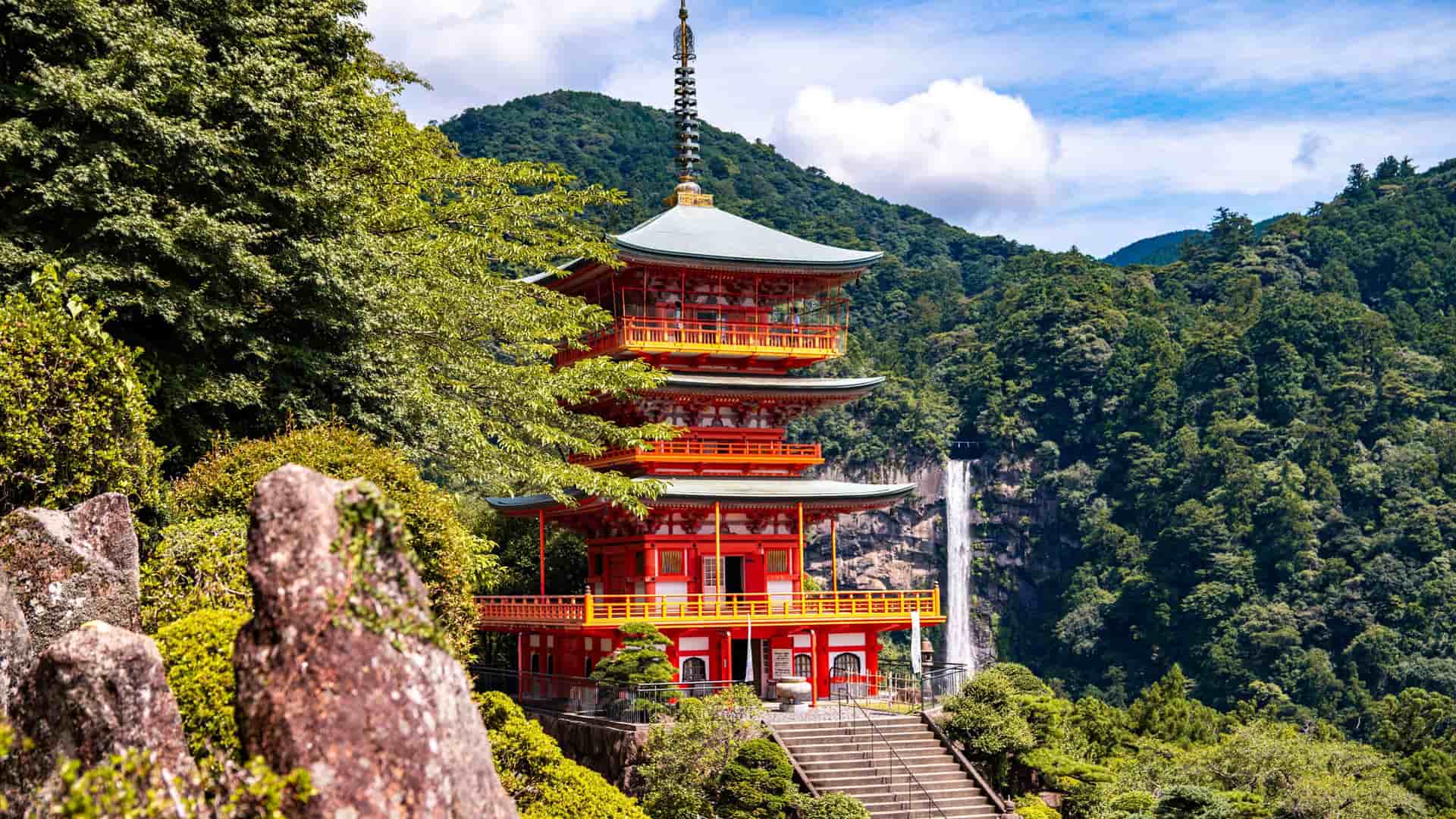 A vibrant red pagoda, the Seiganto-ji Temple, stands majestically amidst lush greenery with the Nachi Falls cascading in the background, a famous landmark near Wakayama, Japan.