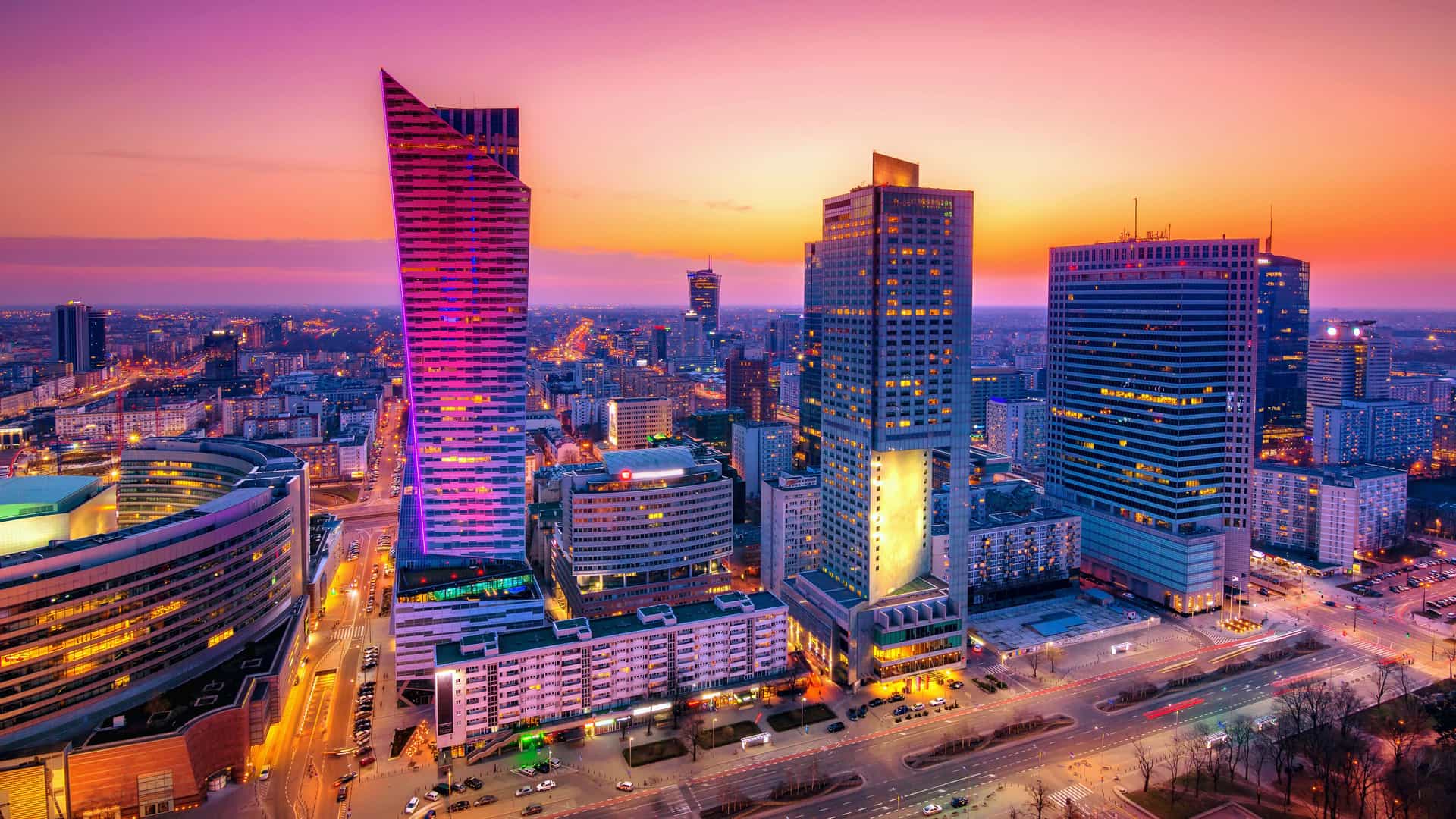 "A stunning panoramic shot of the modern city skyline of Warsaw, Poland, at dusk, with the high-rise buildings illuminated against a vibrant orange and purple sky. "