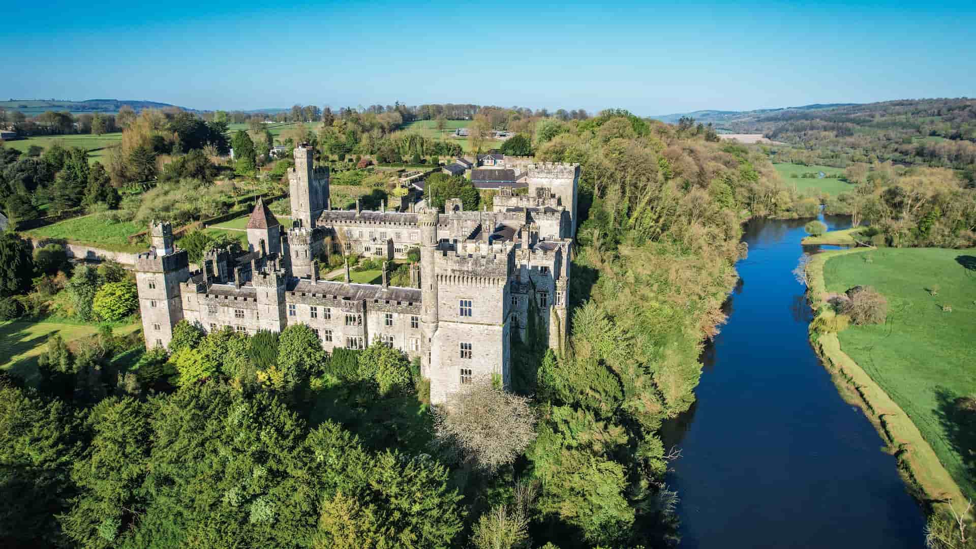 An aerial view of the historic Lismore Castle in County Waterford, Ireland, located on a bank of the River Blackwater and surrounded by lush green forests.