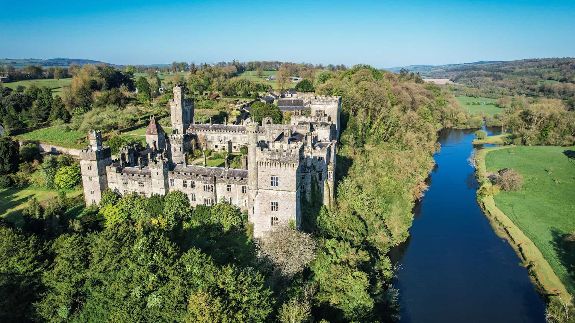 An aerial view of the historic Lismore Castle in County Waterford, Ireland, located on a bank of the River Blackwater and surrounded by lush green forests.
