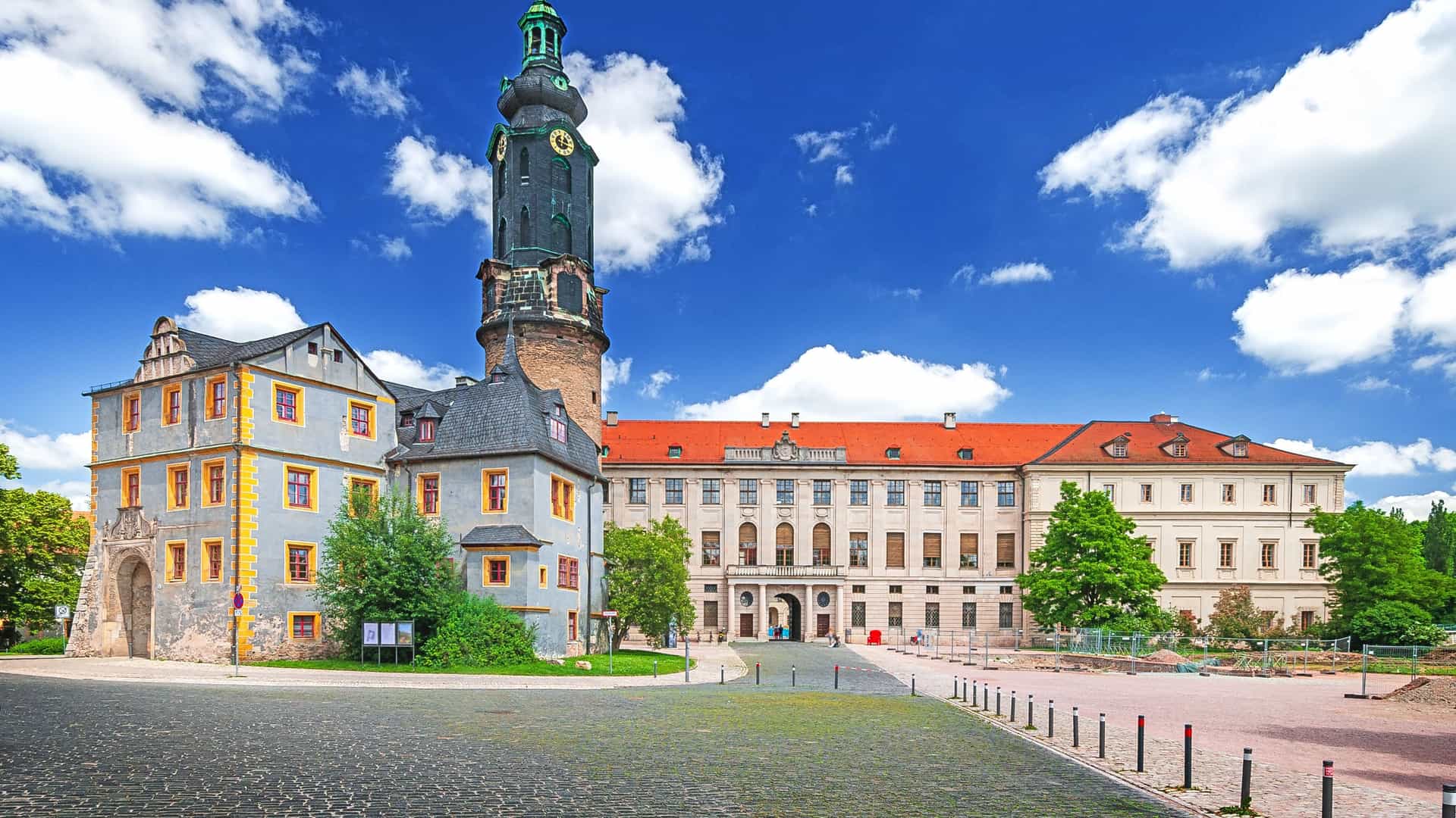 "A sunny, wide-angle shot of the historic Weimar City Palace in Germany, showcasing its large clock tower and Renaissance-style architecture against a vibrant blue sky. "