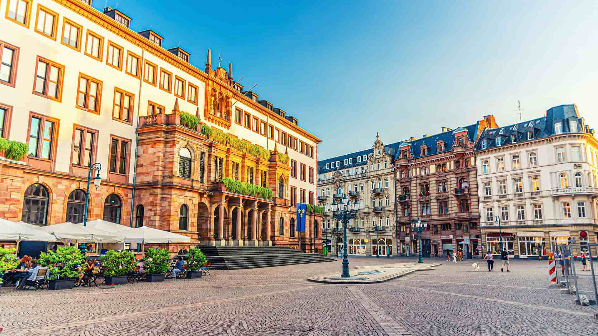 "A vibrant, sunlit view of the historic city center of Wiesbaden, Germany, featuring the grand Old Town Hall with a terraced cafe and colorful buildings in the background. "