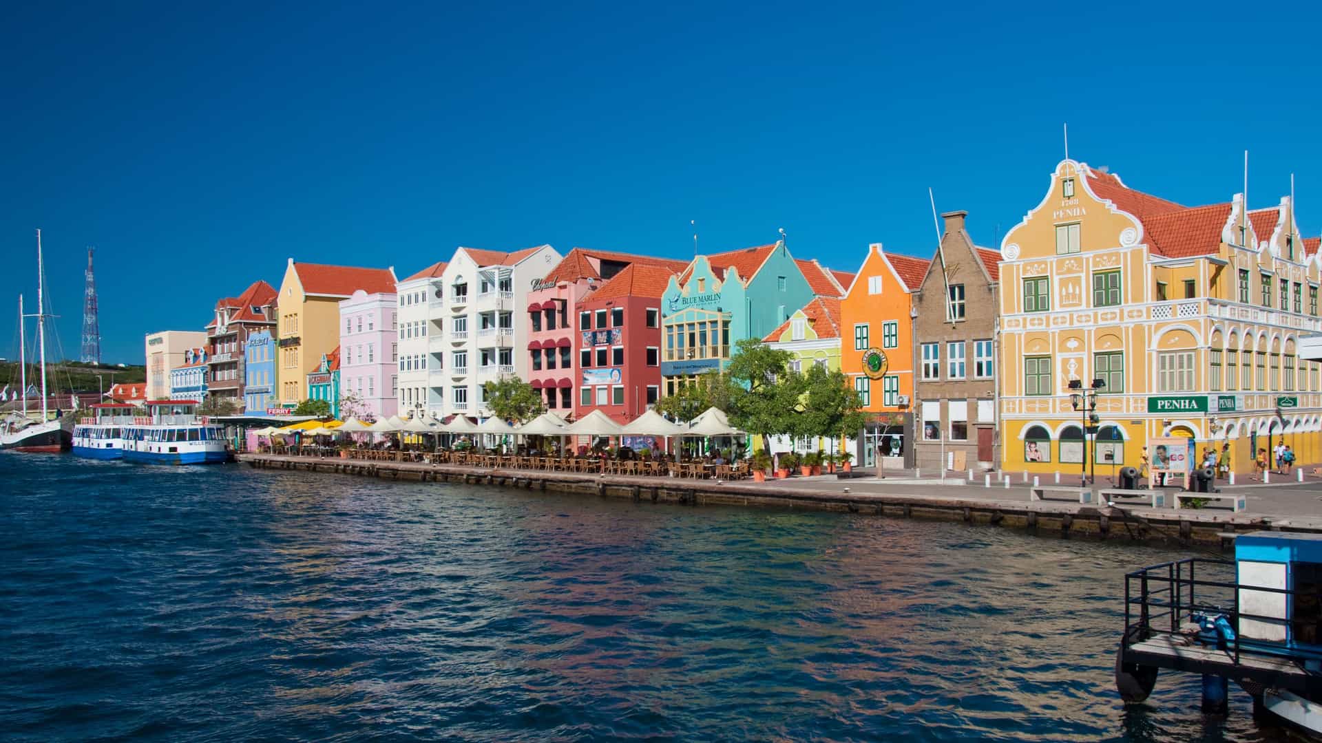 A sunny view of the colorful waterfront of Willemstad, Curacao, with its iconic pastel-colored Dutch colonial buildings lining the Handelskade street and a ship docked in the port.