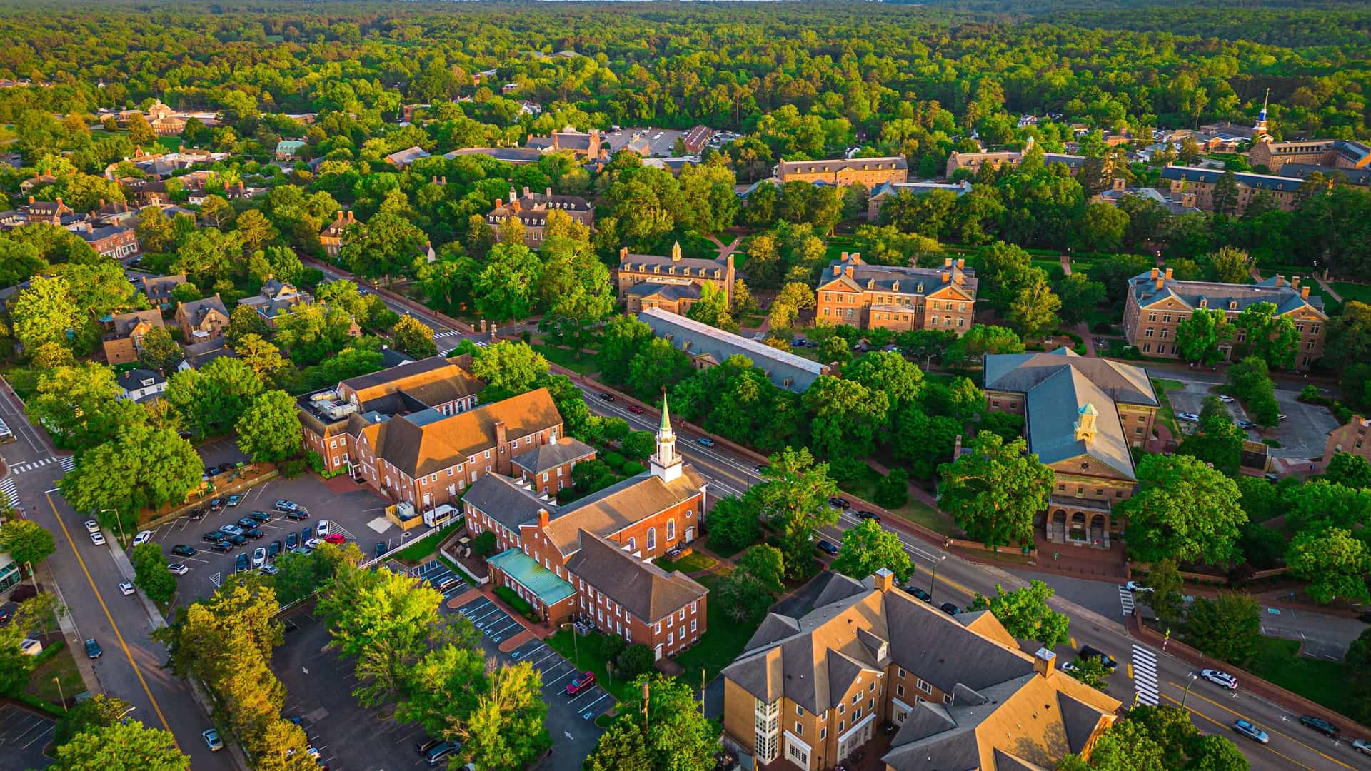 An aerial view of the historic college buildings of William & Mary in Williamsburg, Virginia, surrounded by a dense green forest stretching to the horizon.