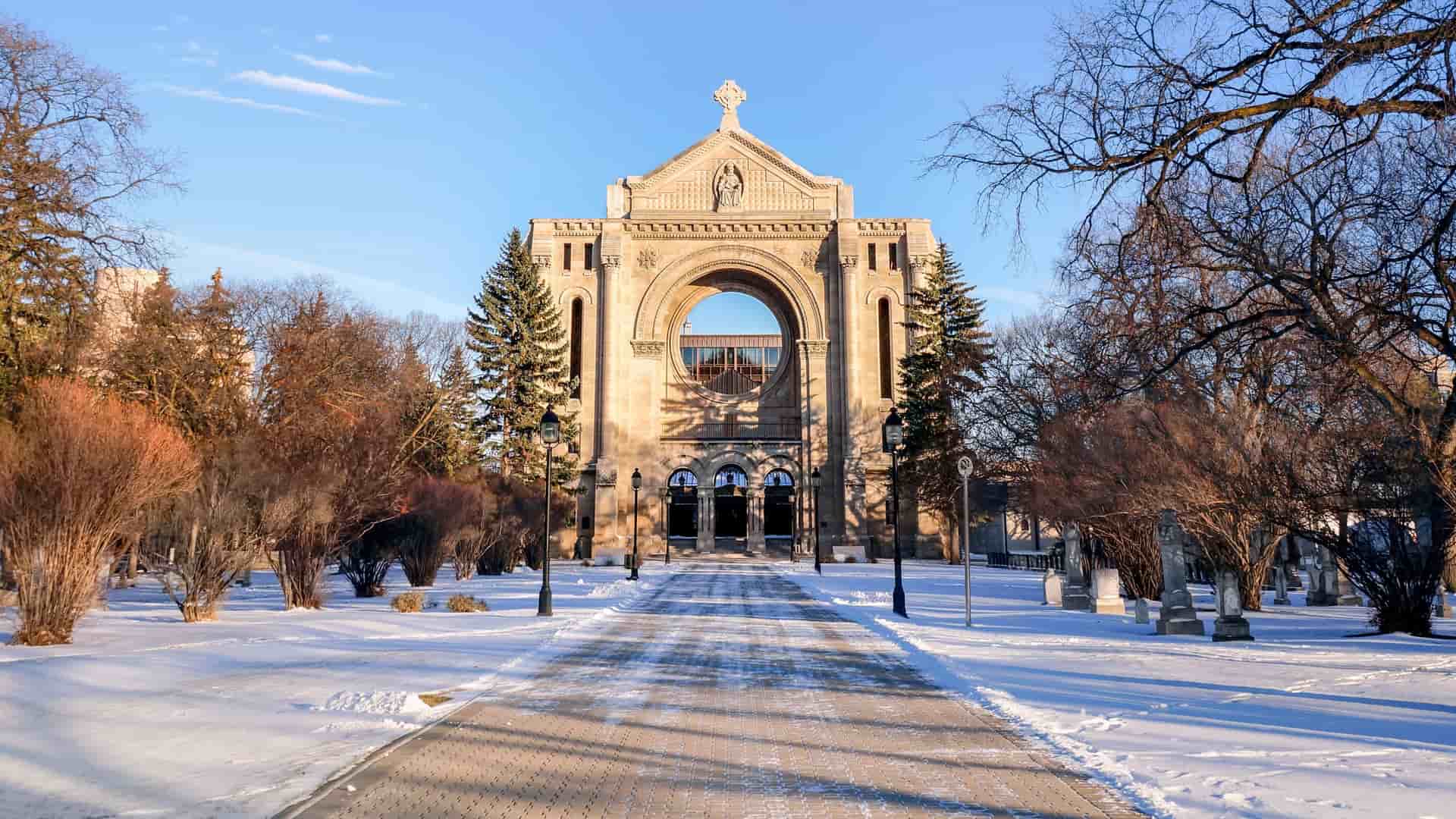 "A stunning winter view of the historic St. Boniface Cathedral-Basilica in Winnipeg, Canada, with a wide, snow-covered pathway leading to the iconic stone facade. "