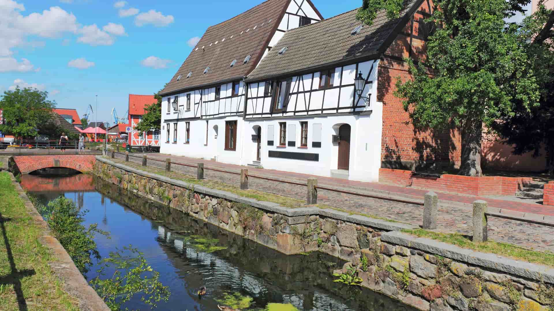 A view of the historic Old Town of Wismar, Germany, with a peaceful canal and half-timbered houses lining the cobblestone street under a sunny sky.