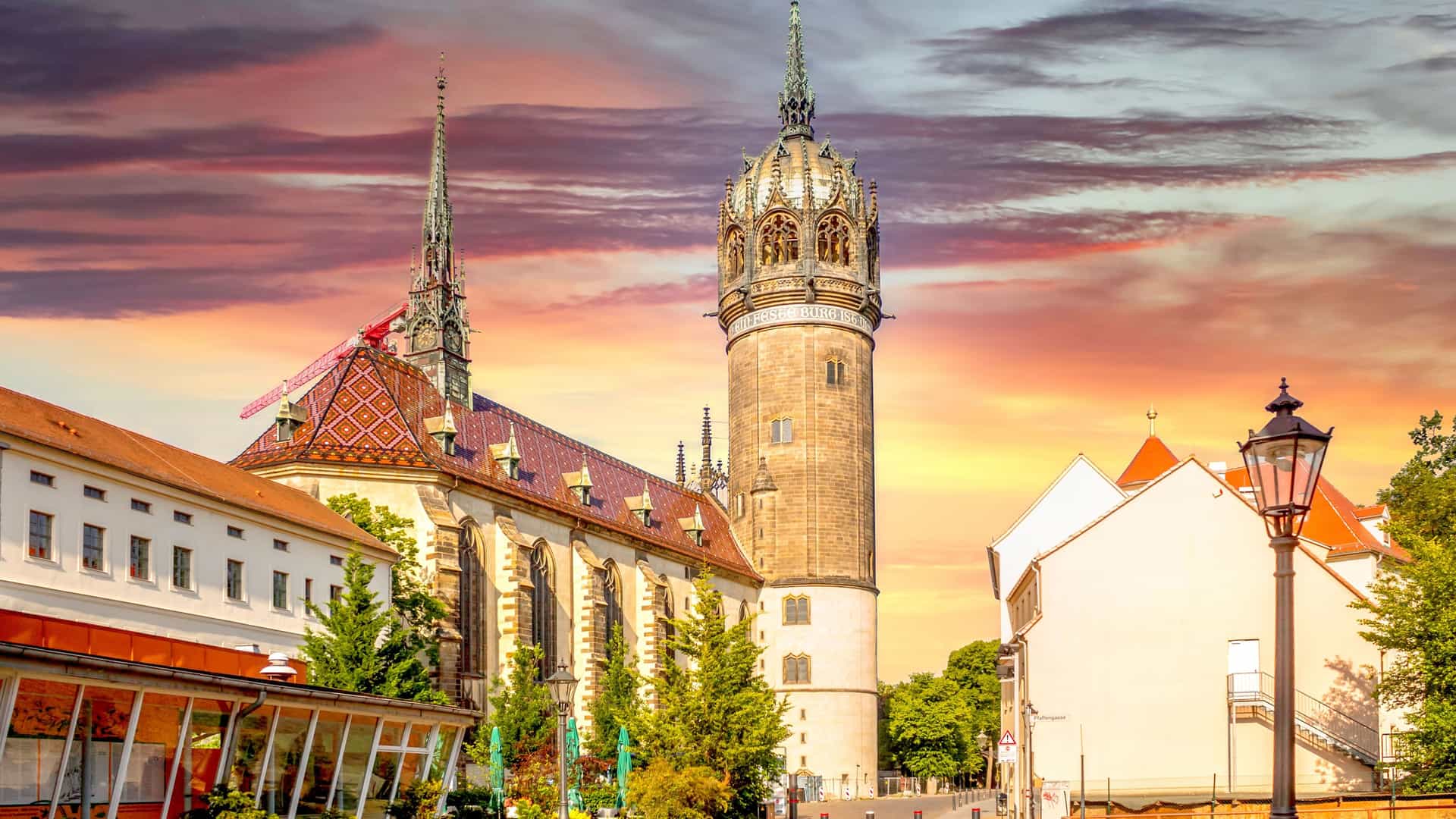 "A breathtaking sunset over the historic Wittenberg All Saints' Church in Germany, with its ornate tower and spires against a sky filled with dramatic pink and orange clouds. "