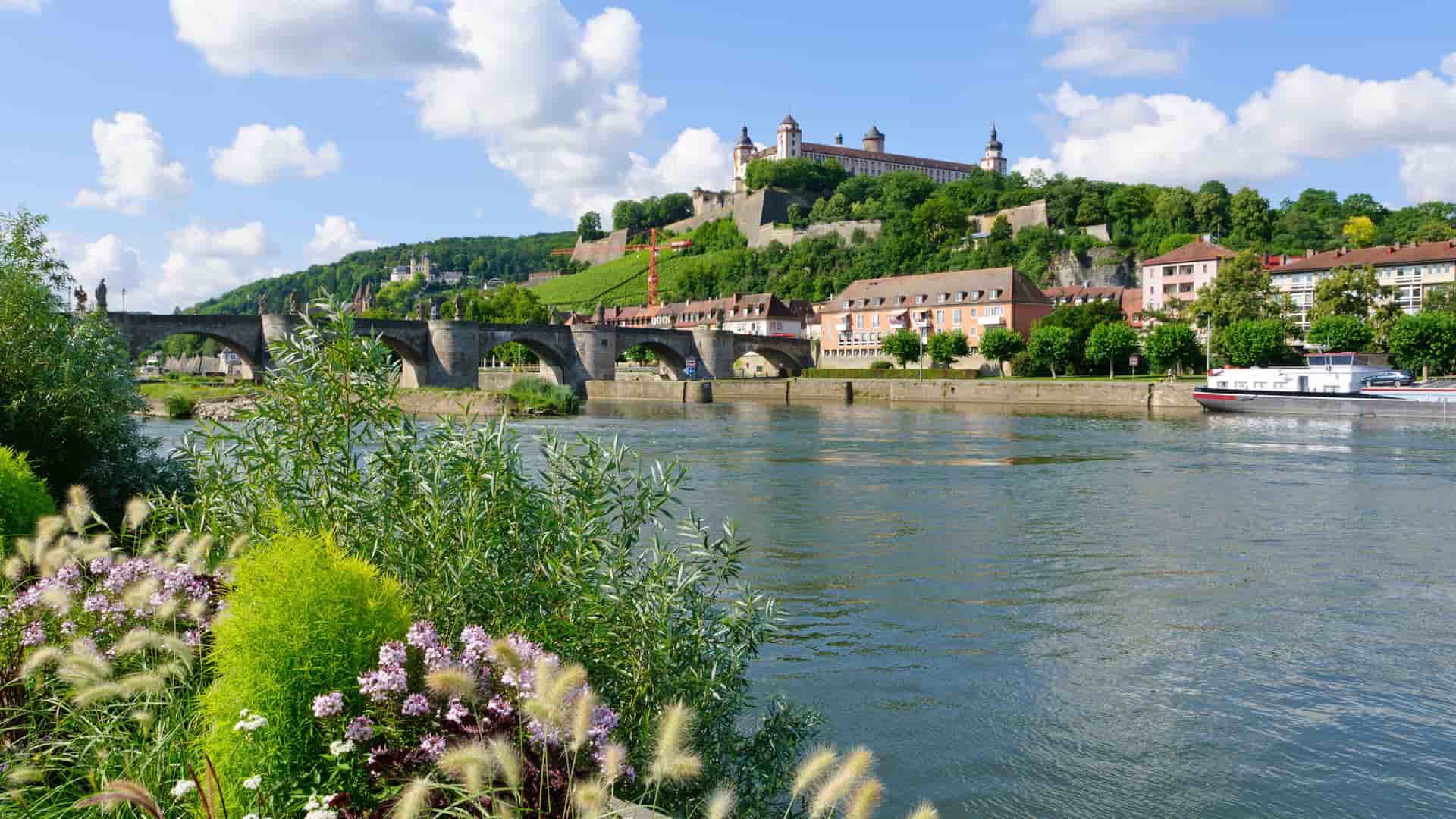 A picturesque view of the Marienberg Fortress towering over the Main River in Würzburg, Germany, with the Old Main Bridge and a river cruise ship.