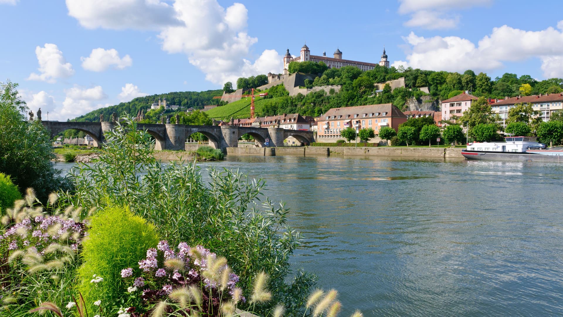 A picturesque view of the Marienberg Fortress towering over the Main River in Würzburg, Germany, with the Old Main Bridge and a river cruise ship.