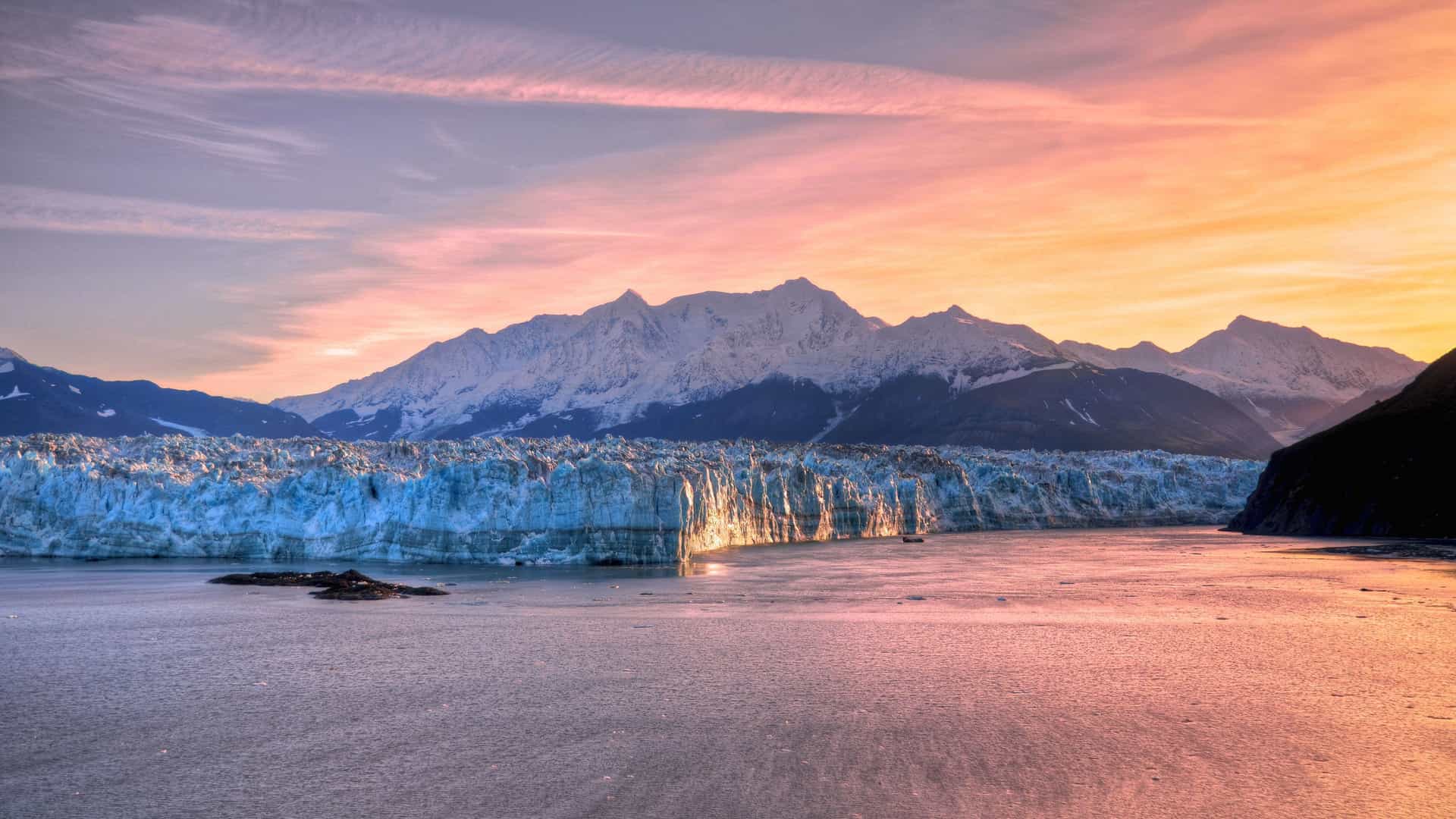 A panoramic view of the Hubbard Glacier in Yakutat Bay, Alaska, with a massive wall of blue ice calving into the water under a dramatic sunset sky.
