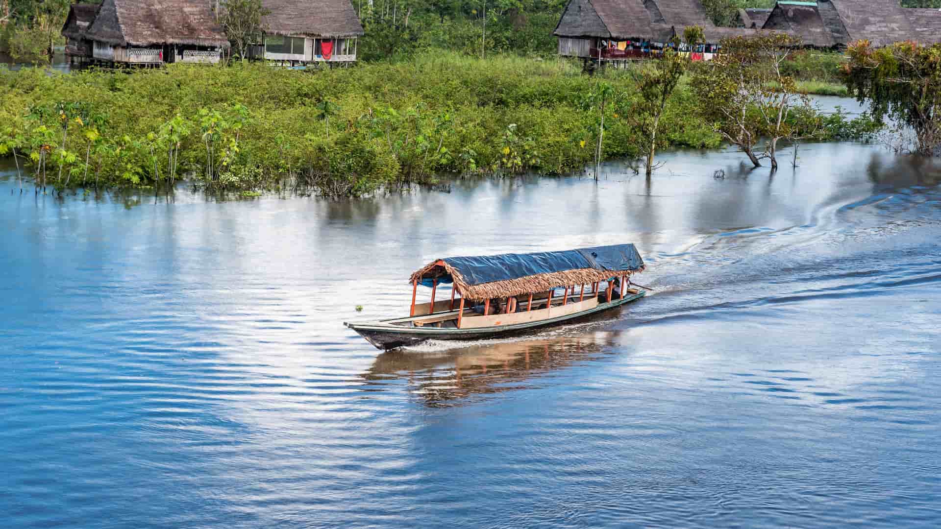 A traditional thatched-roof longboat on the Yarapa River in the Amazon Jungle of Peru, with stilted houses and lush green vegetation visible on the riverbank in the background.