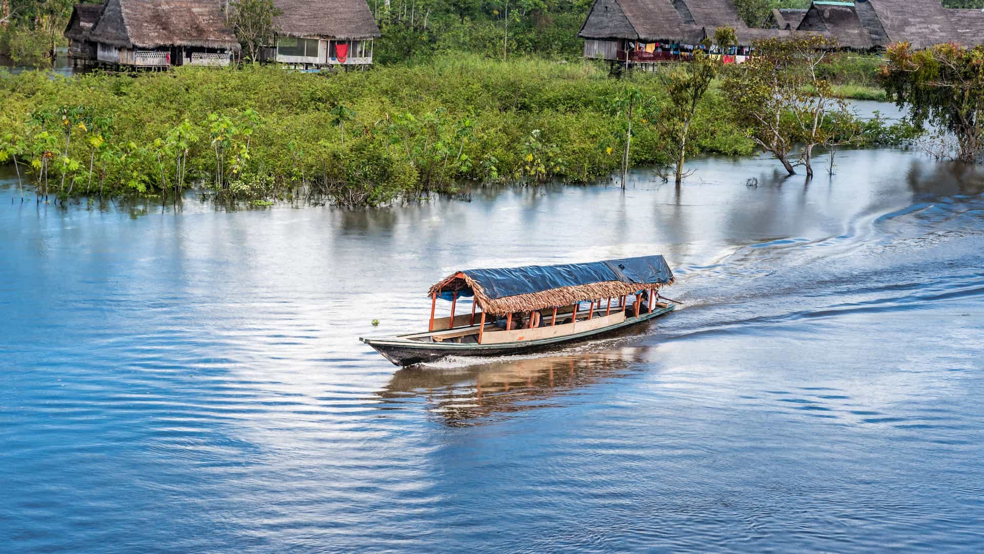A traditional thatched-roof longboat on the Yarapa River in the Amazon Jungle of Peru, with stilted houses and lush green vegetation visible on the riverbank in the background.