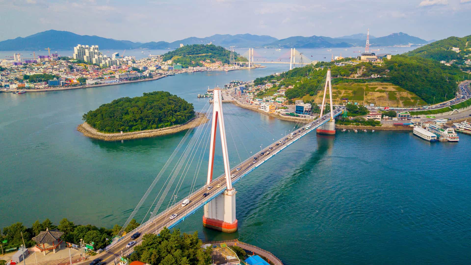 An aerial view of the Dolsan Bridge, a modern cable-stayed bridge in Yeosu, South Korea, connecting Dolsan Island to the mainland with a city and mountains in the background.