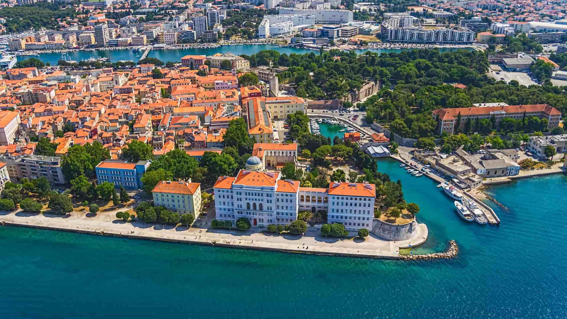 An aerial view of Zadar, Croatia, showcasing the historic old town with its red-tiled roofs, the marina filled with boats, and the stunning blue waters of the Adriatic Sea.