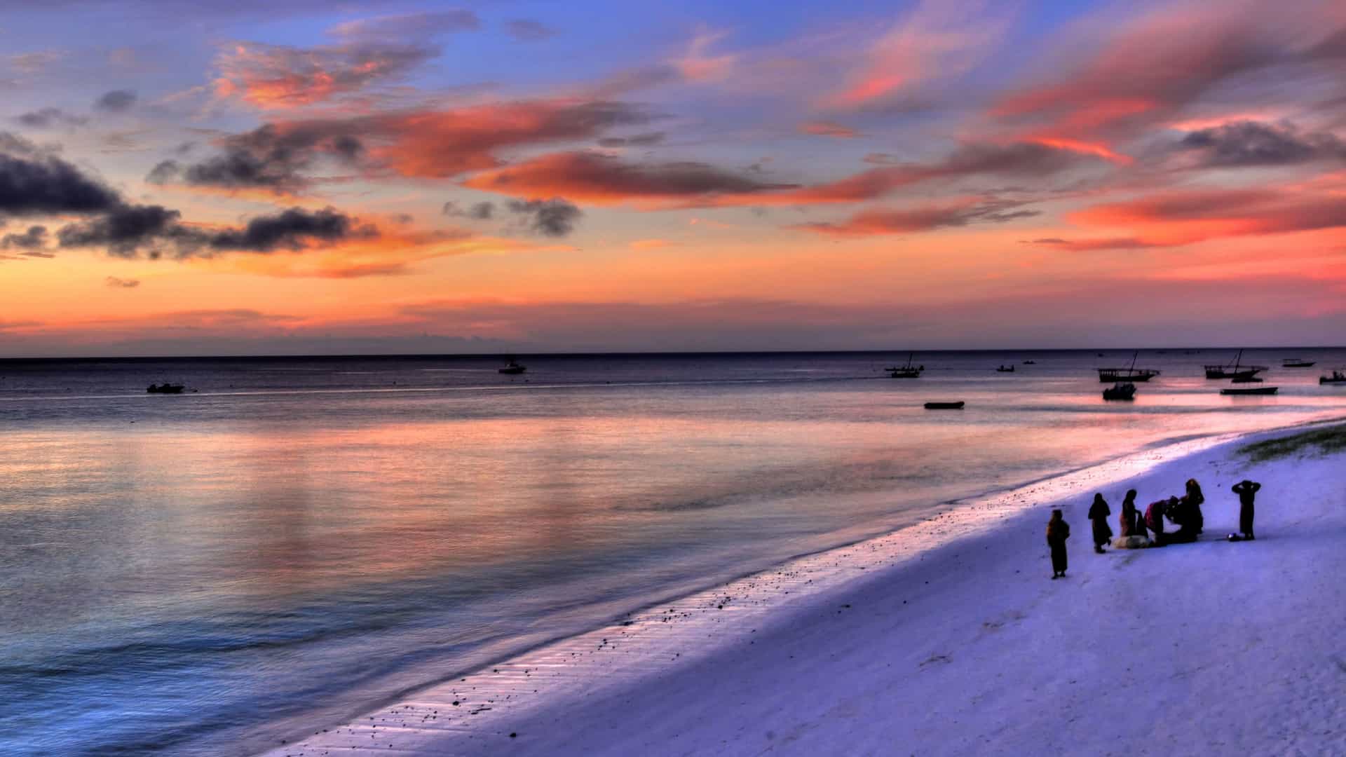 A stunning multi-colored sunset over the coast of Zanzibar, with traditional fishing boats silhouetted on the calm ocean and people gathered on the white sand beach.