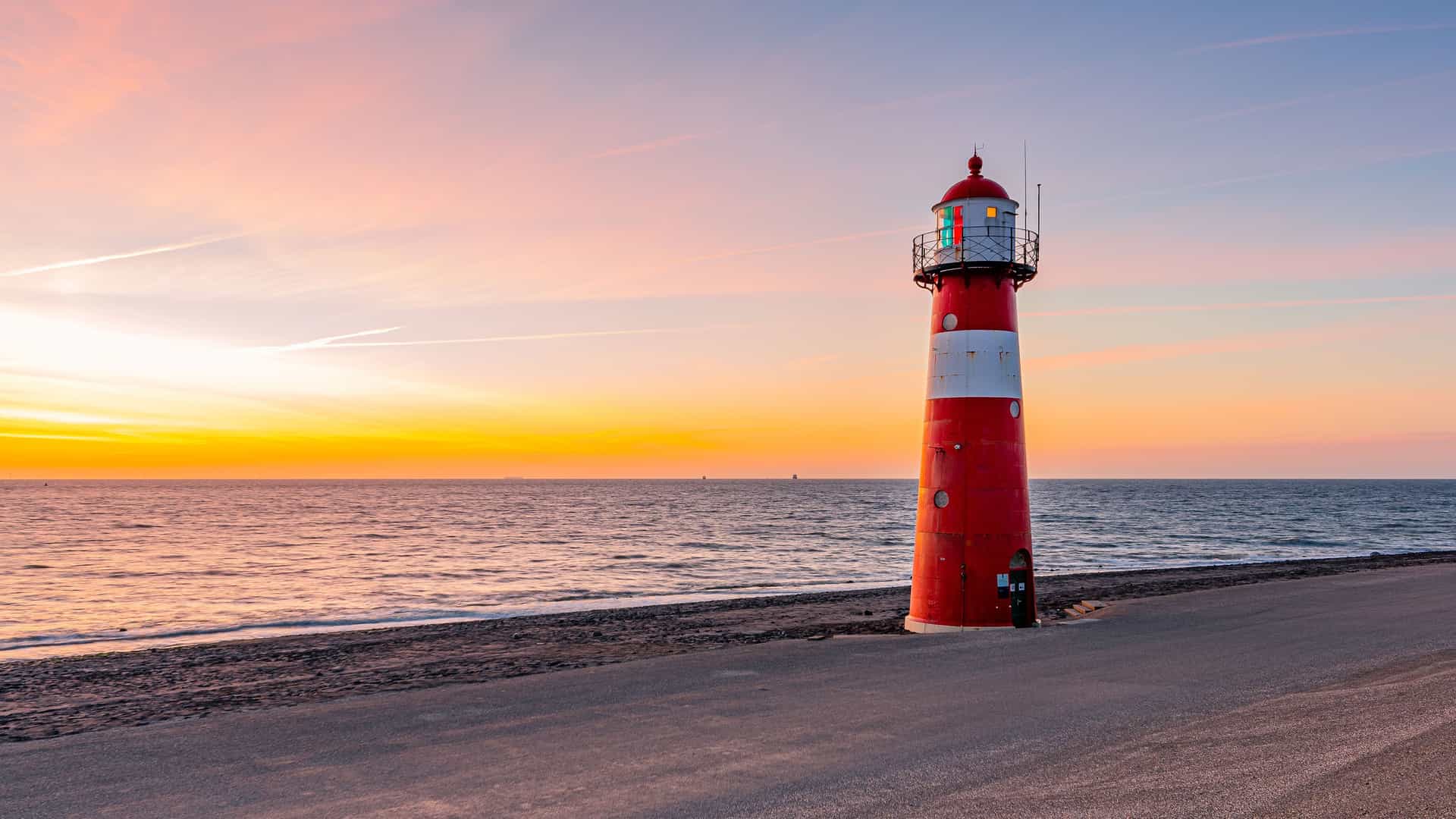 A stunning horizontal shot of the red and white lighthouse of Westkapelle in Zeeland, Netherlands, against a beautiful orange and blue sunset over the North Sea.